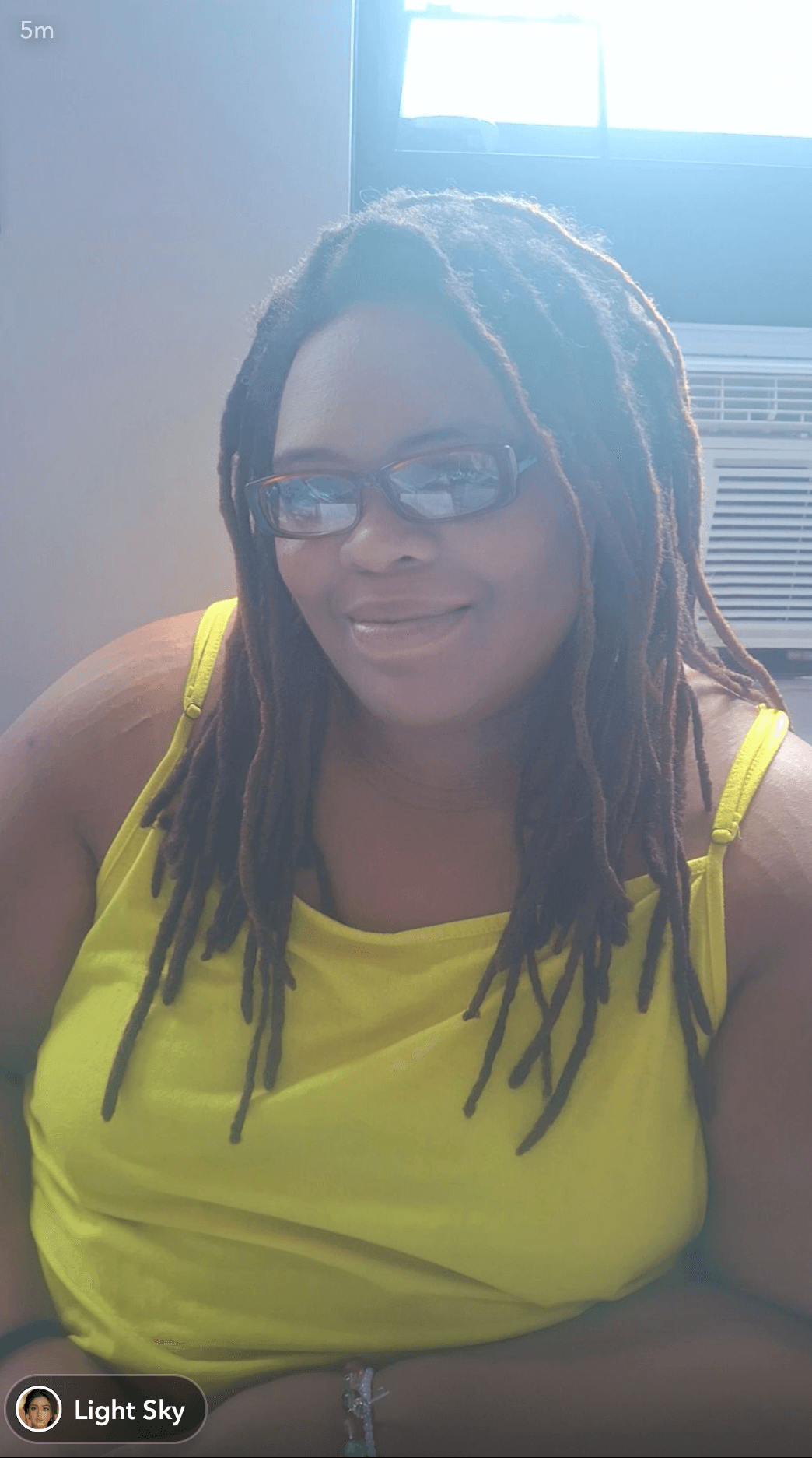 Woman with glasses and dreadlocks wearing a yellow top, sitting indoors near a window.