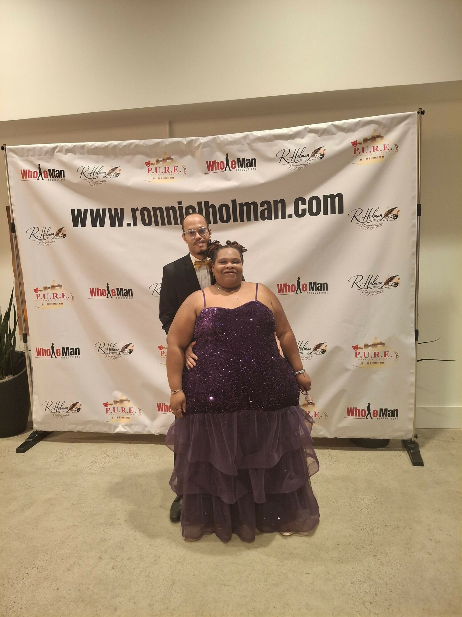 Woman in a purple gown standing in front of a promotional backdrop, holding a phone, with a beige floor and wall.