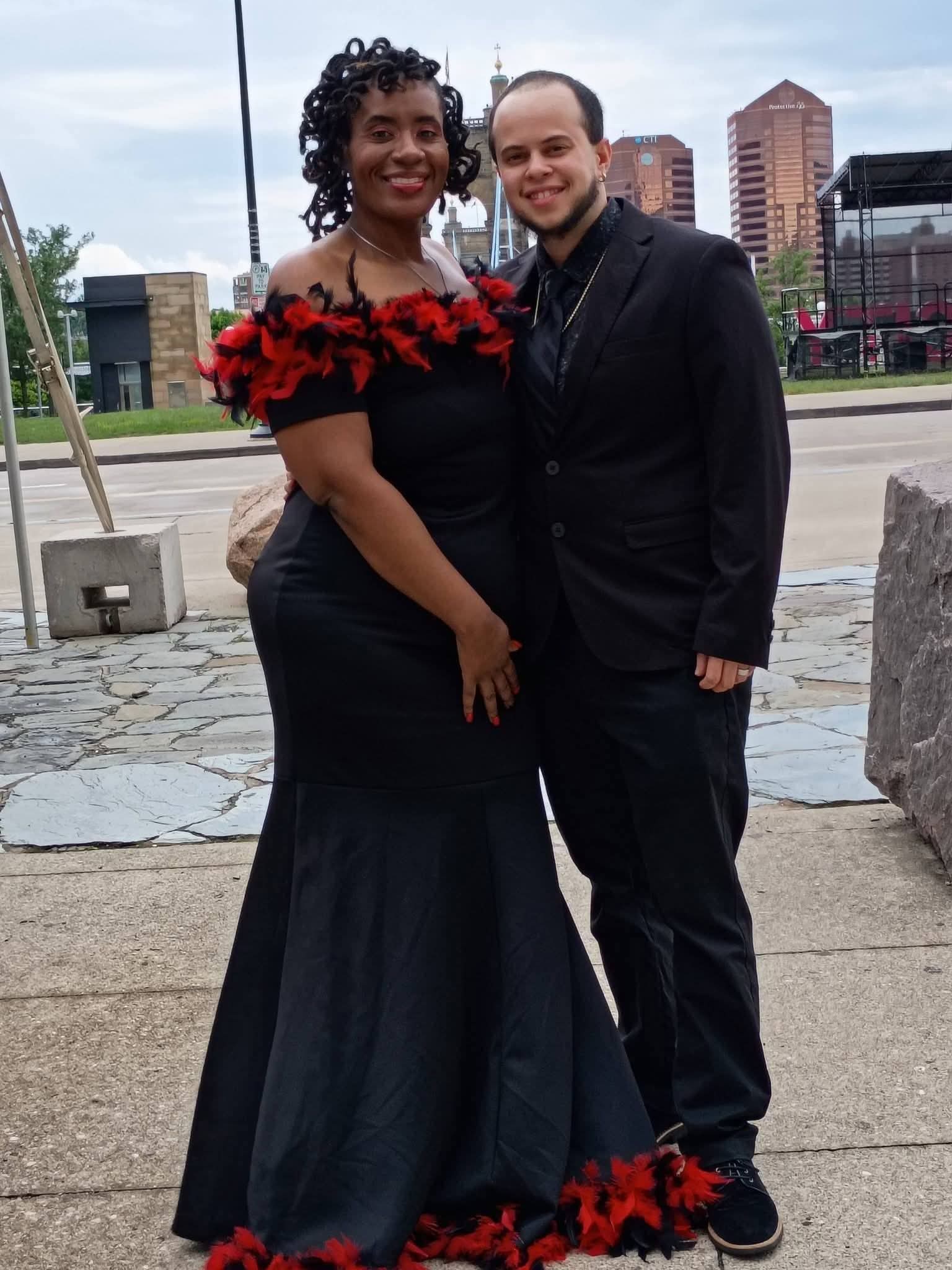 Two people standing outdoors, smiling, with city buildings in the background, one in a black dress with red floral details, the other in a black suit.