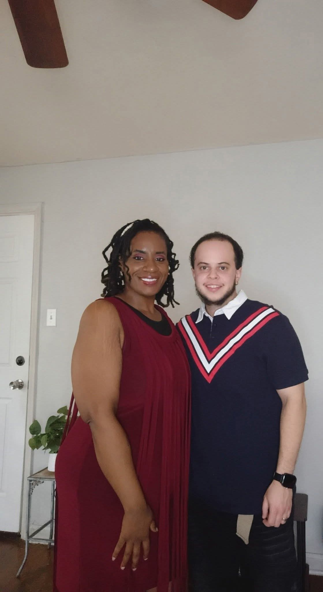 Two people standing indoors, one in a red dress and the other in a navy sweater with red and white stripes, smiling.