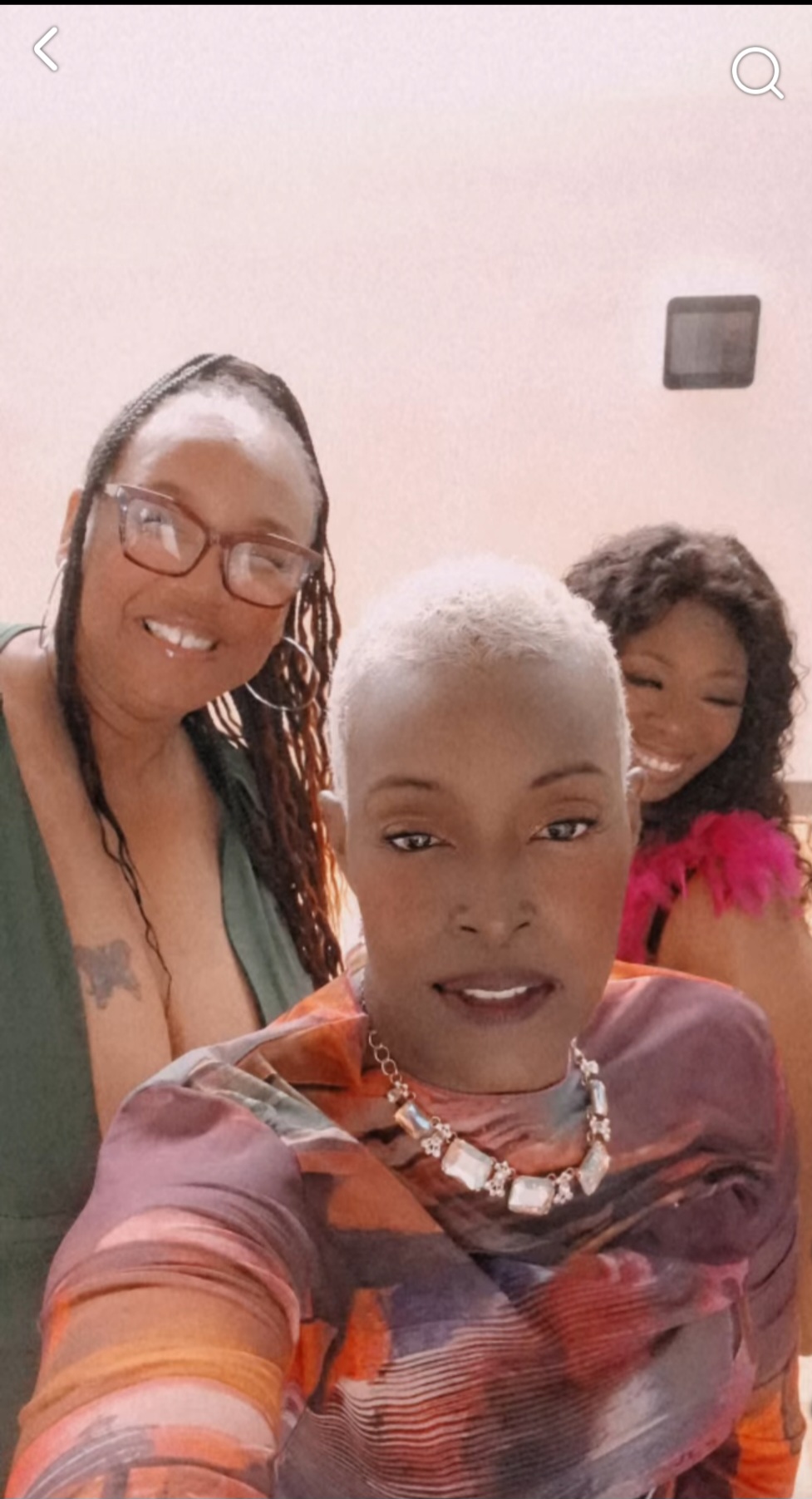 Three women smiling, one with short white hair and glasses, another with long dark hair, and a third with curly hair, indoors.