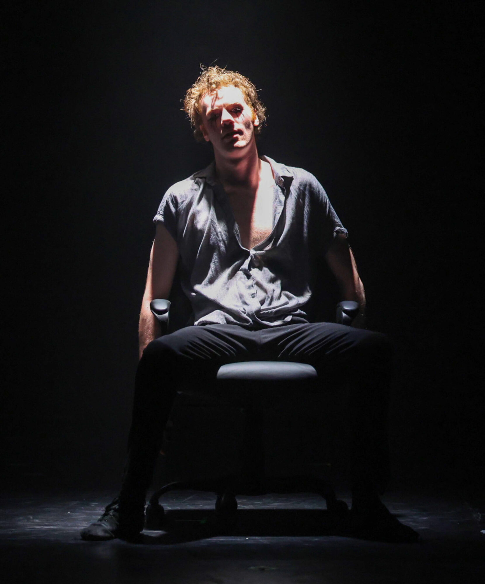 Person with curly hair sitting on a chair in a dark setting, illuminated by a spotlight, with a serious expression.