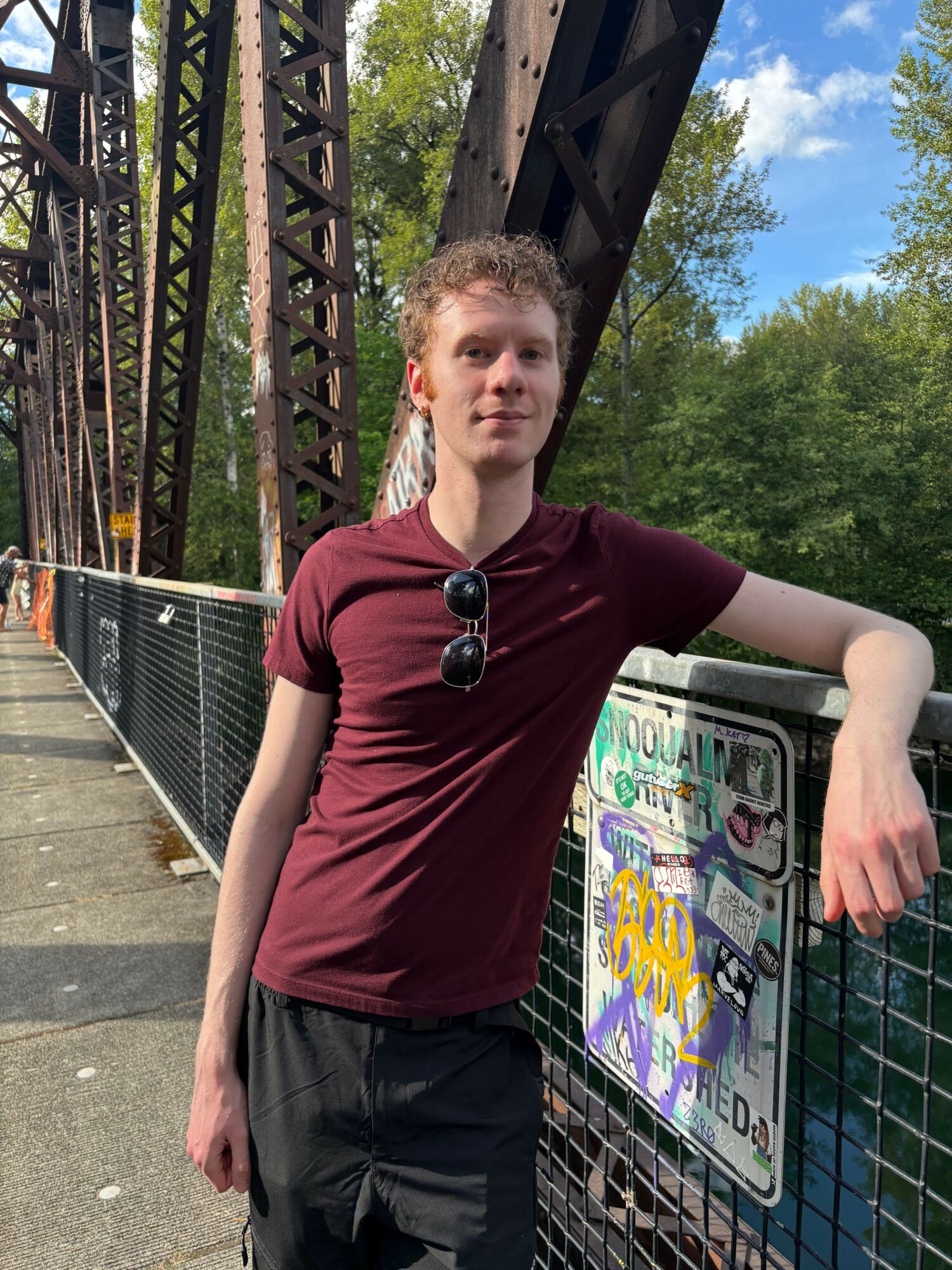 Young man with curly hair wearing a maroon t-shirt and sunglasses hanging from his shirt, standing outdoors near a metal fence and a colorful sign, with trees and blue sky in the background.