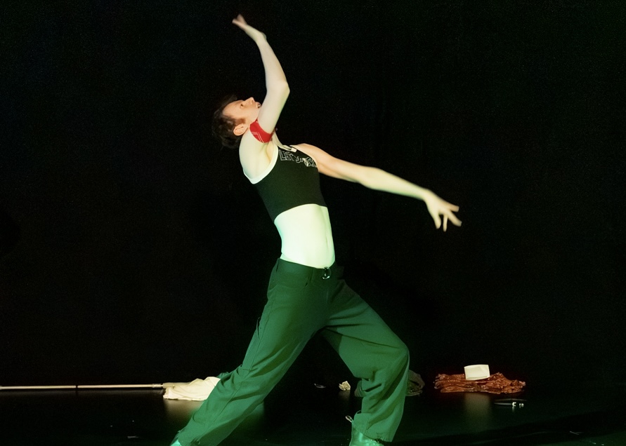Person performing a dance move with one arm raised and the other extended sideways, wearing green pants and a black top.