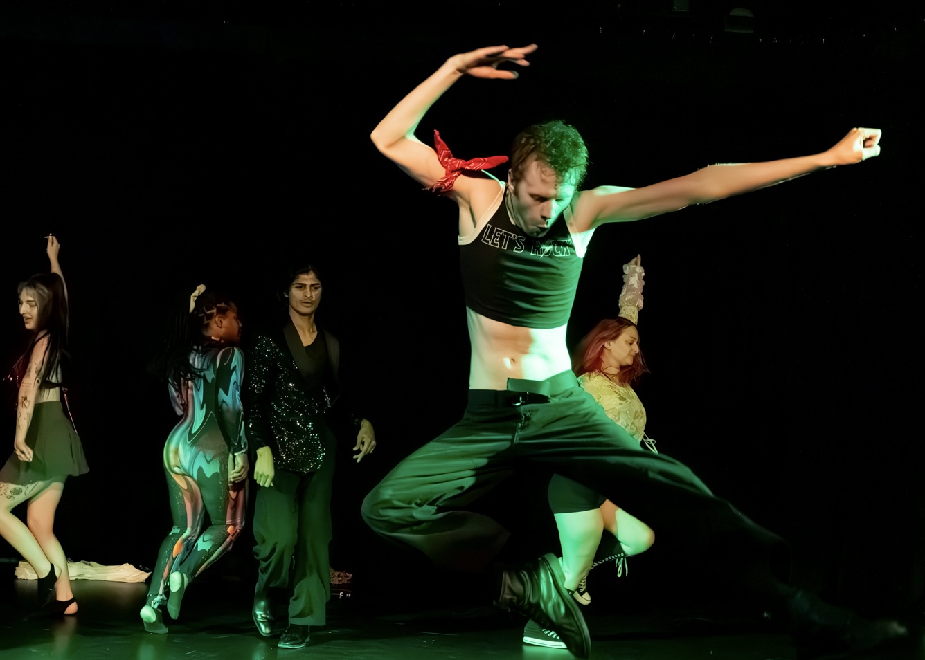 A dancer jumps with arms extended, wearing a black and red outfit, on stage with other performers in the background.