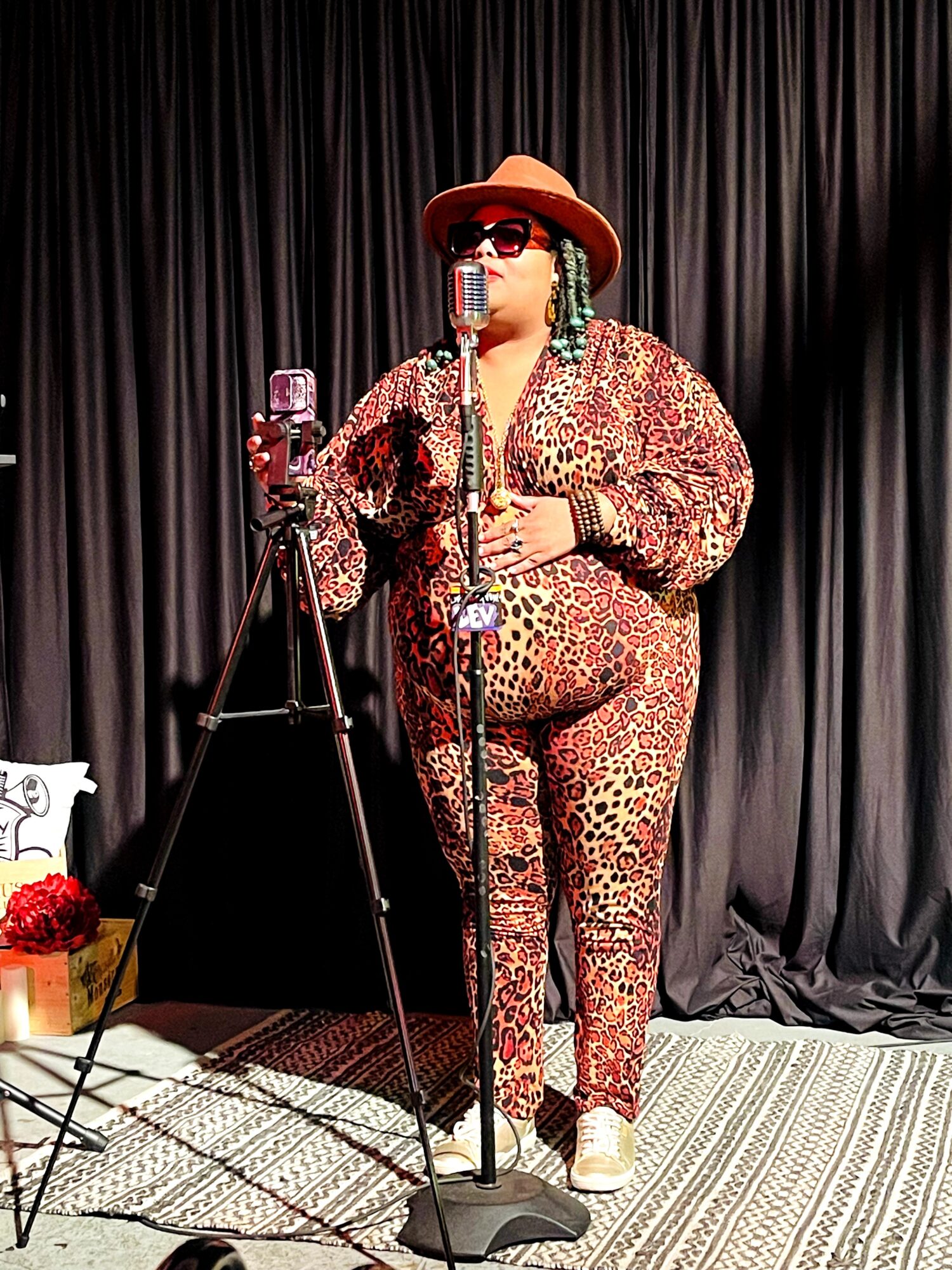 A person in a leopard-print outfit and brown hat stands at a microphone beside a camera on a tripod.