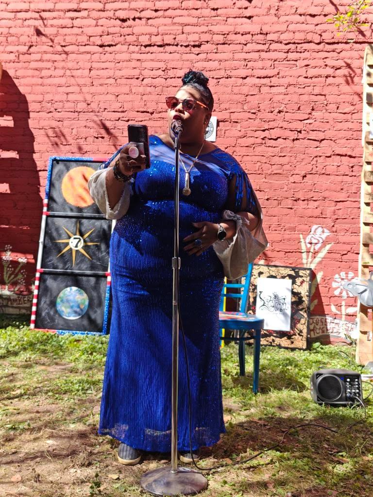 Person in a blue dress singing into a microphone outdoors against a painted brick wall.