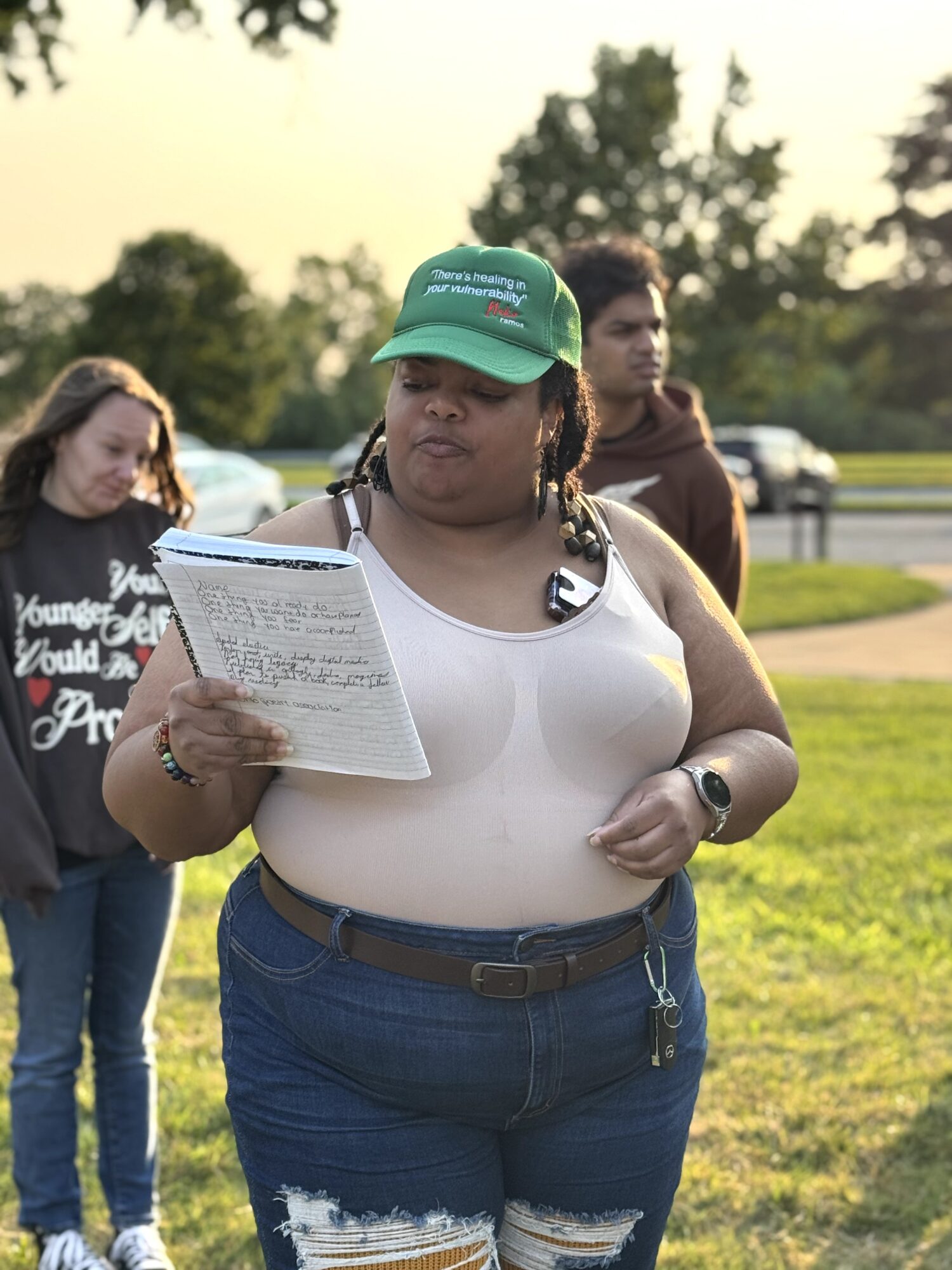 Person in a green cap reads a sheet of paper outdoors, with two people standing behind.