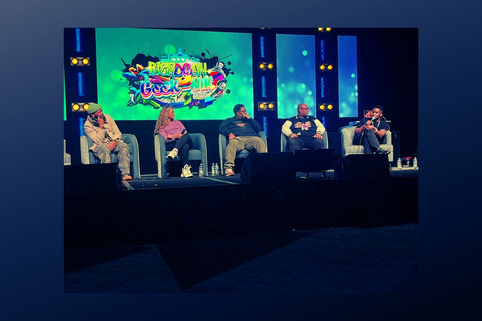 Five people sit on a stage with a colorful screen behind them, engaged in a discussion or panel. Stage lighting is visible.