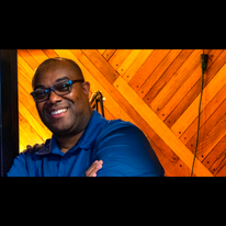 Smiling man with glasses in a blue shirt, wooden wall background, arms crossed, indoors.