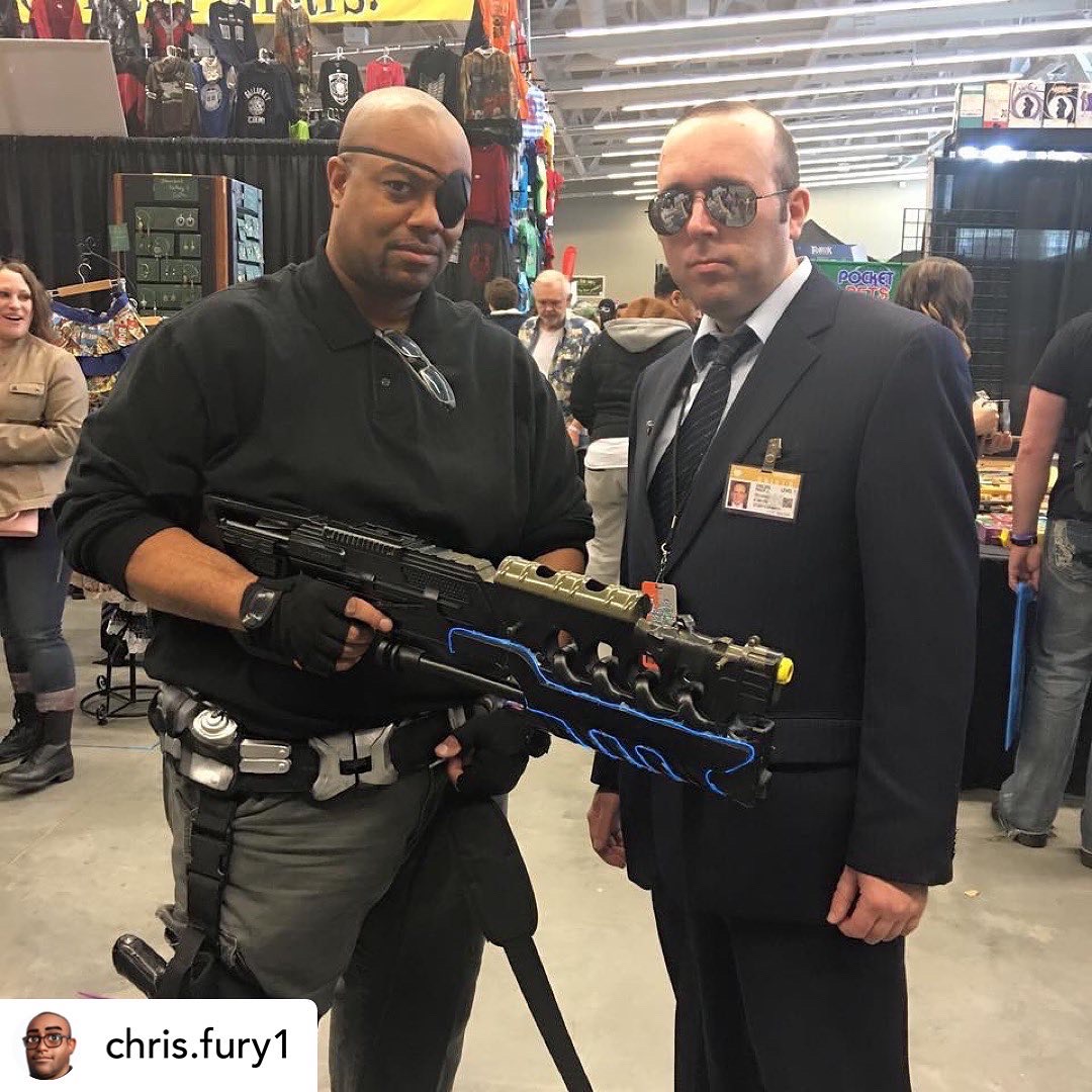 Two men stand indoors, one in tactical gear holding a large gun, the other in a suit with sunglasses, surrounded by people.