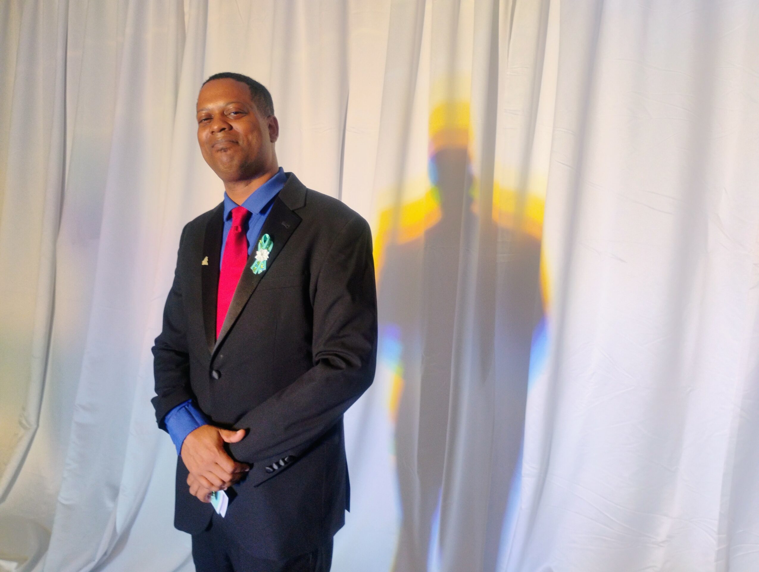 Man in a suit standing in front of a white curtain with a colorful shadow behind him.