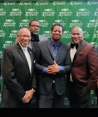 Four men in formal attire standing in front of a green backdrop with white text, smiling and posing for a photo.