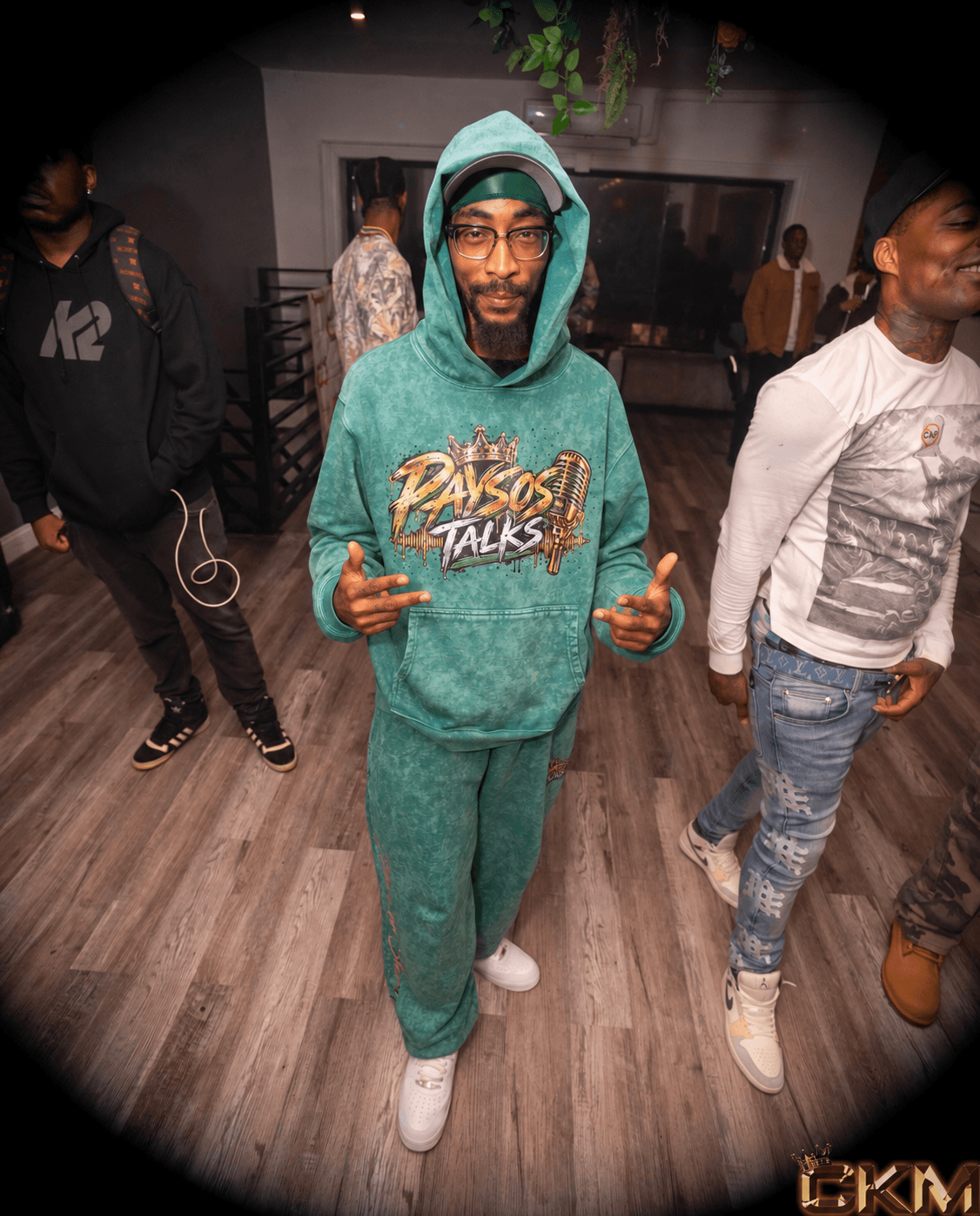 Man in teal hoodie and sweatpants making hand gestures at a social gathering, with other people in the background.