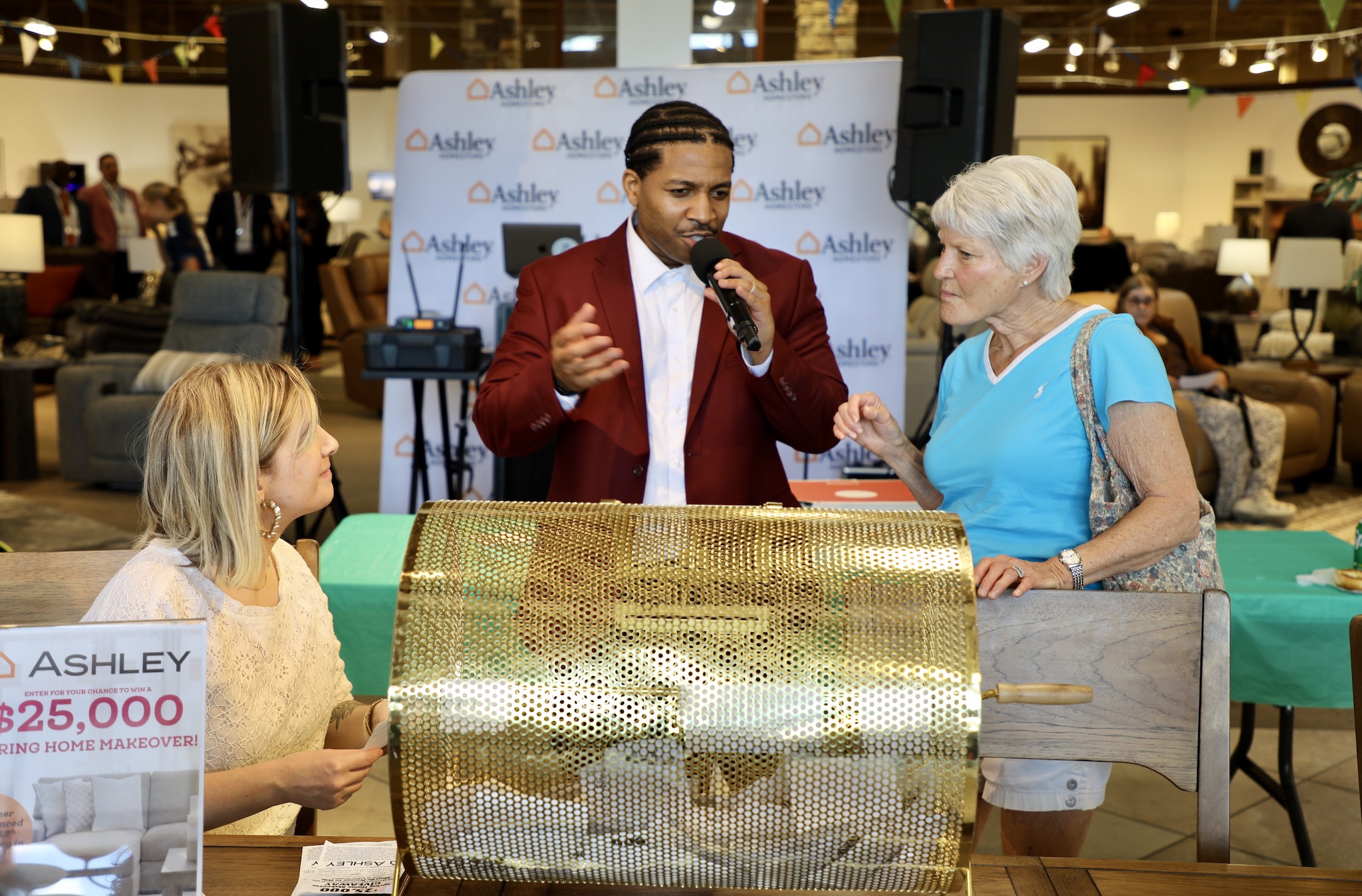Man in red blazer speaks into microphone while woman in blue listens, with a young girl nearby, at an event booth.