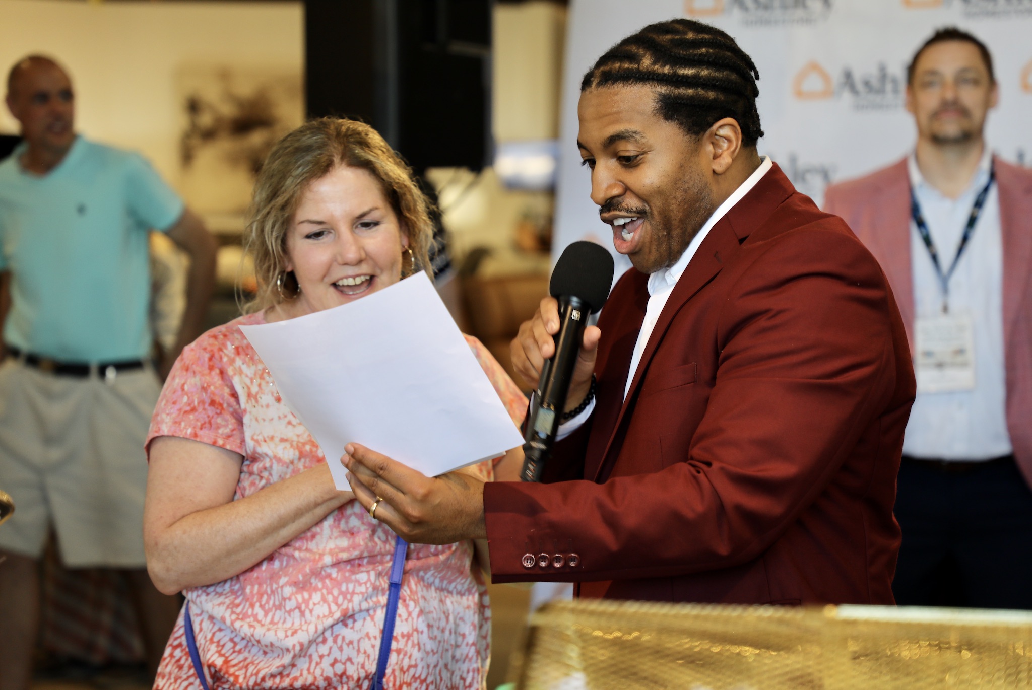 Man in red suit holding microphone and paper, smiling, woman in pink shirt reading paper, people in background.
