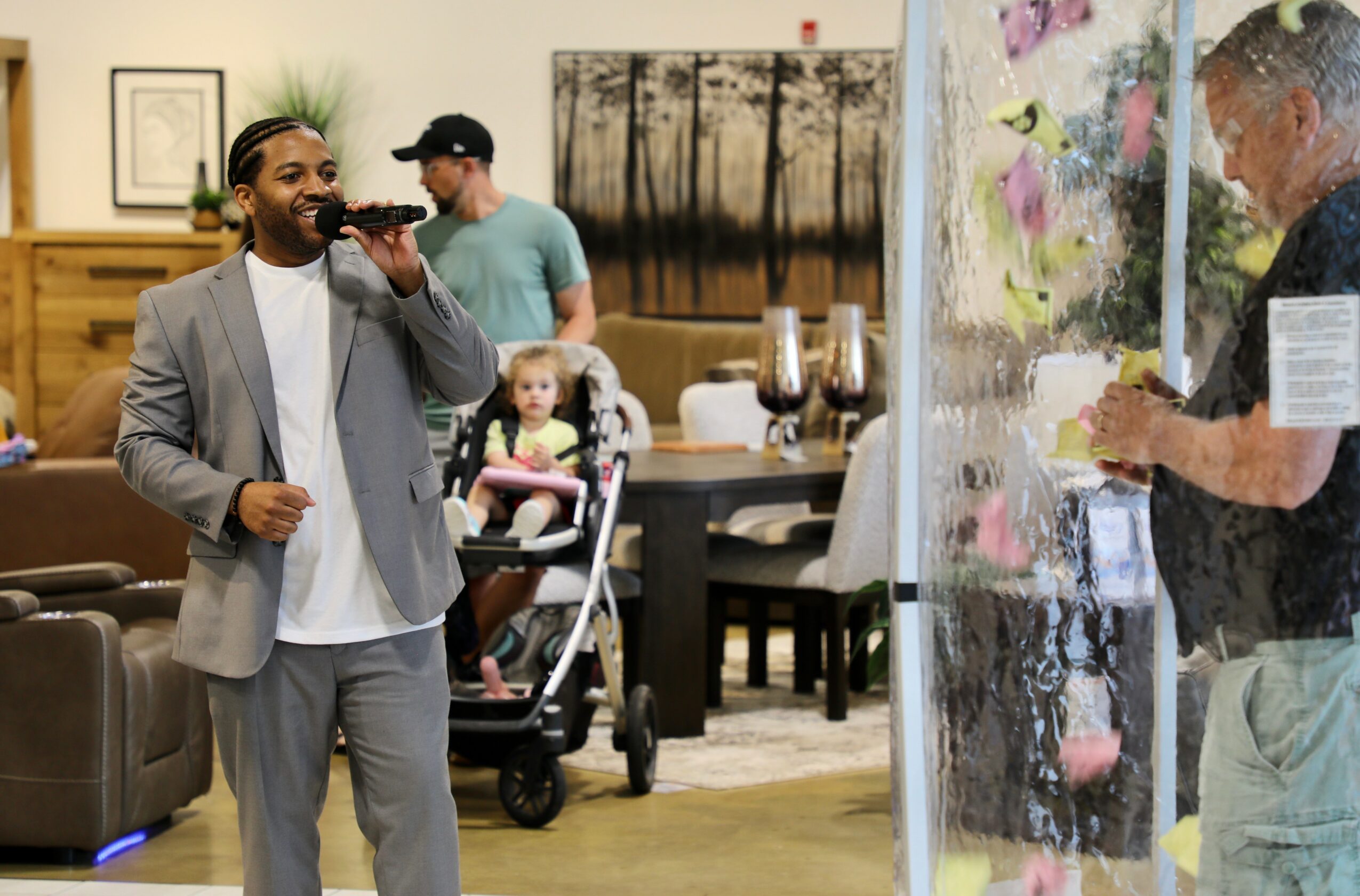 Man in gray suit talking on phone in a room with people, a child in stroller, and artwork on walls.