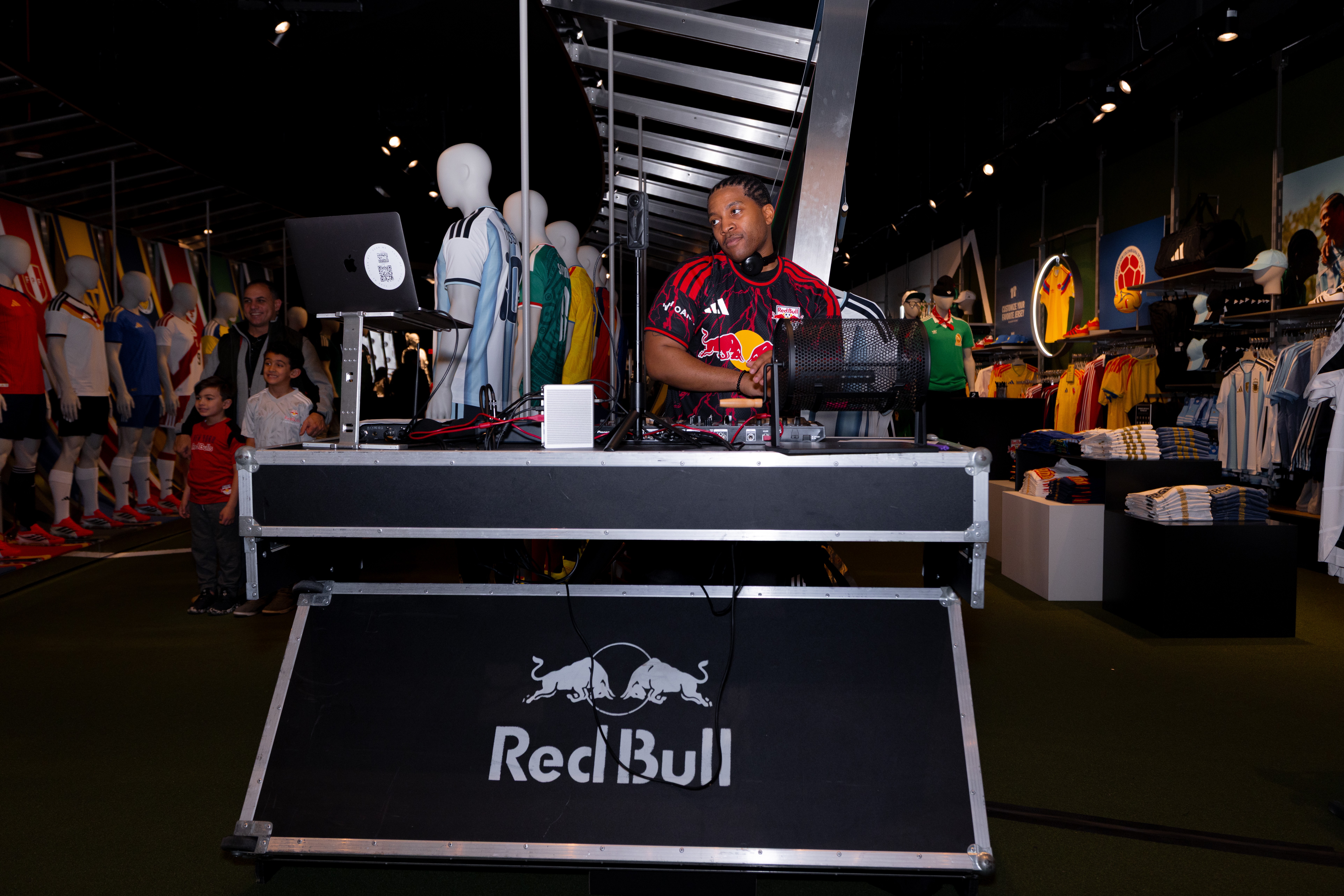 A person in a Red Bull sports jersey stands behind a table at an indoor event, with clothing displays and people in the background.