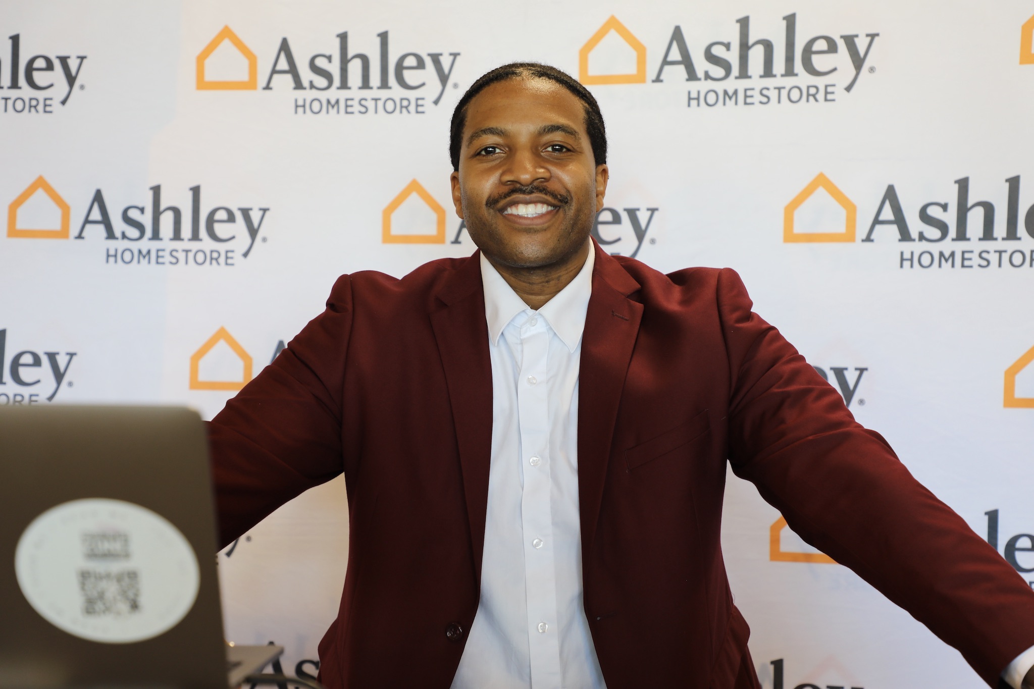 Man in a red blazer smiling with arms outstretched in front of a backdrop with 'Ashley HomeStore' logos.
