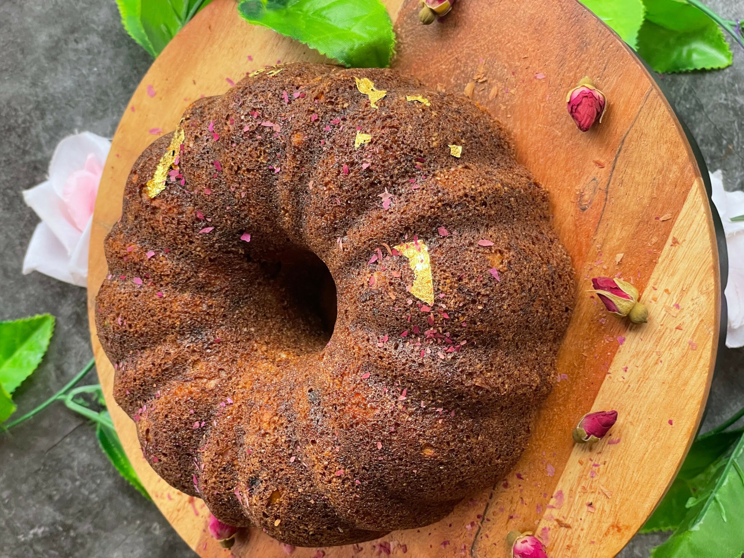 Bundt cake on a wooden board with pink rosebuds and green leaves around it.