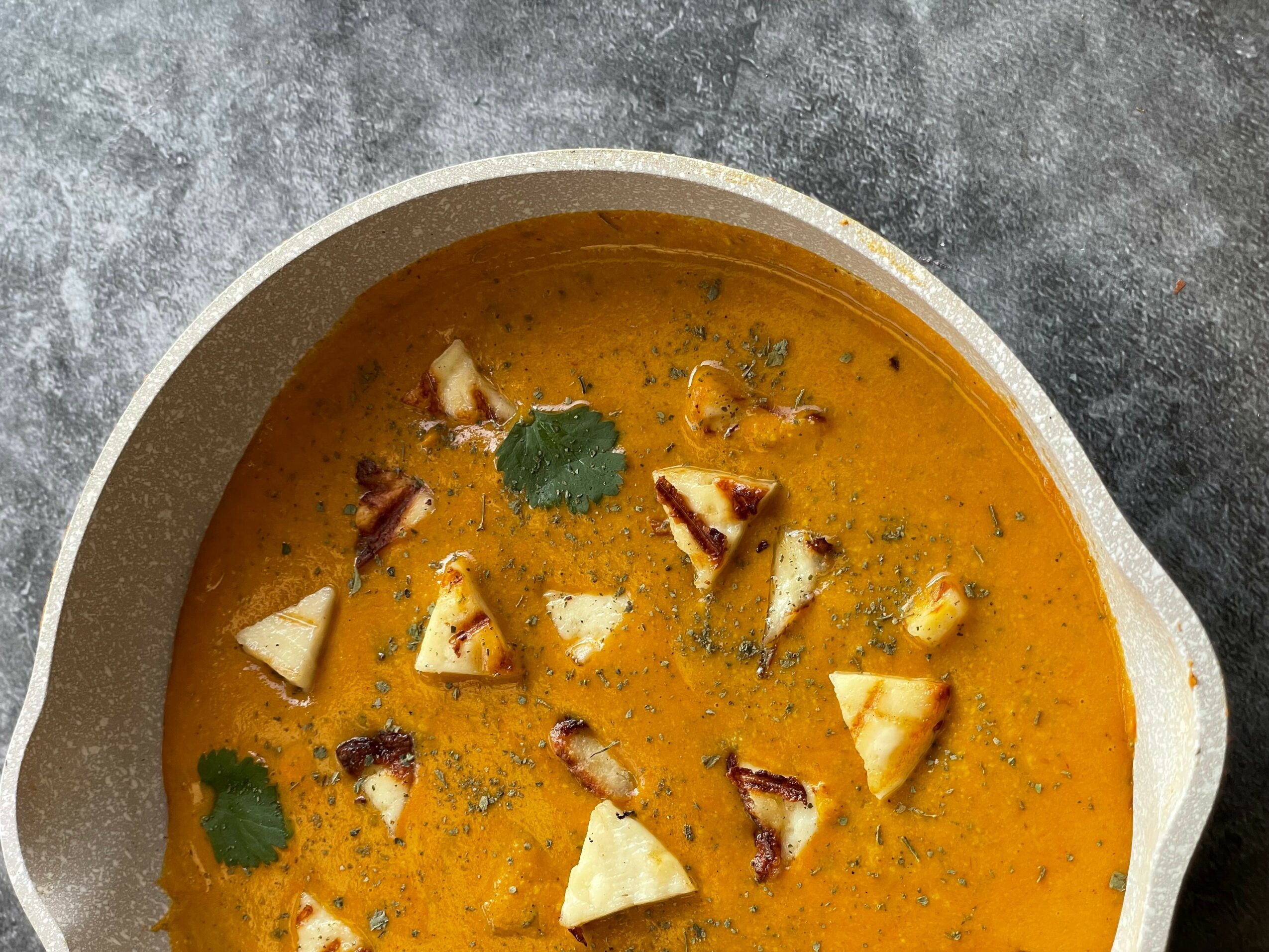 Bowl of orange-colored soup garnished with chopped vegetables and herbs, on a gray surface.