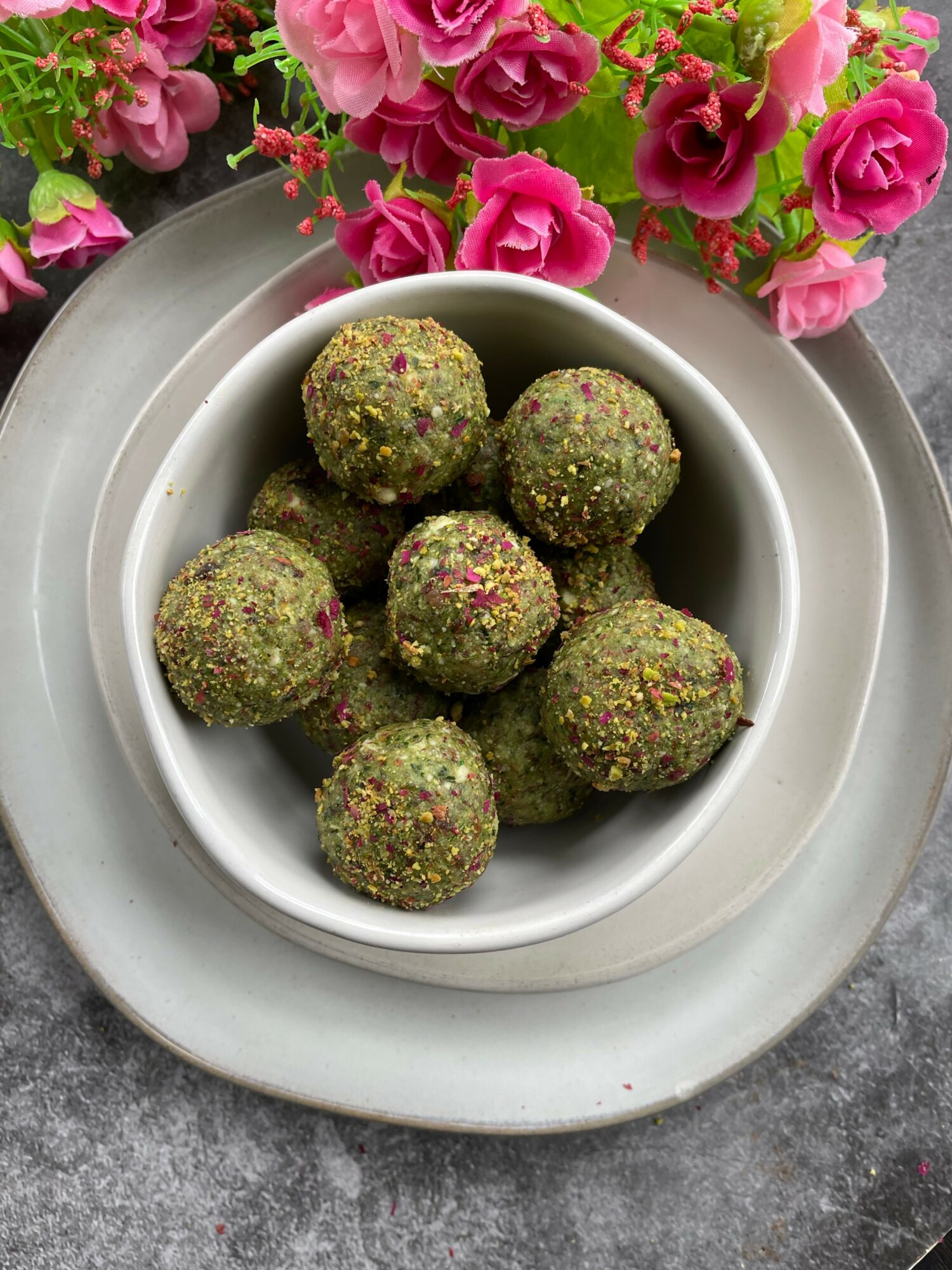 Bowl of green round snacks with colorful sprinkles, placed on a white plate with pink flowers in background.