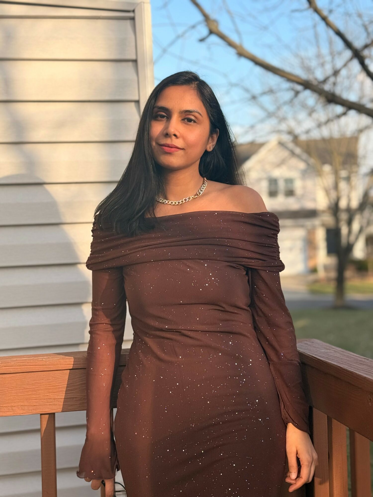 Young woman with long dark hair wearing an off-shoulder brown dress and necklace, standing outdoors near a house and trees.
