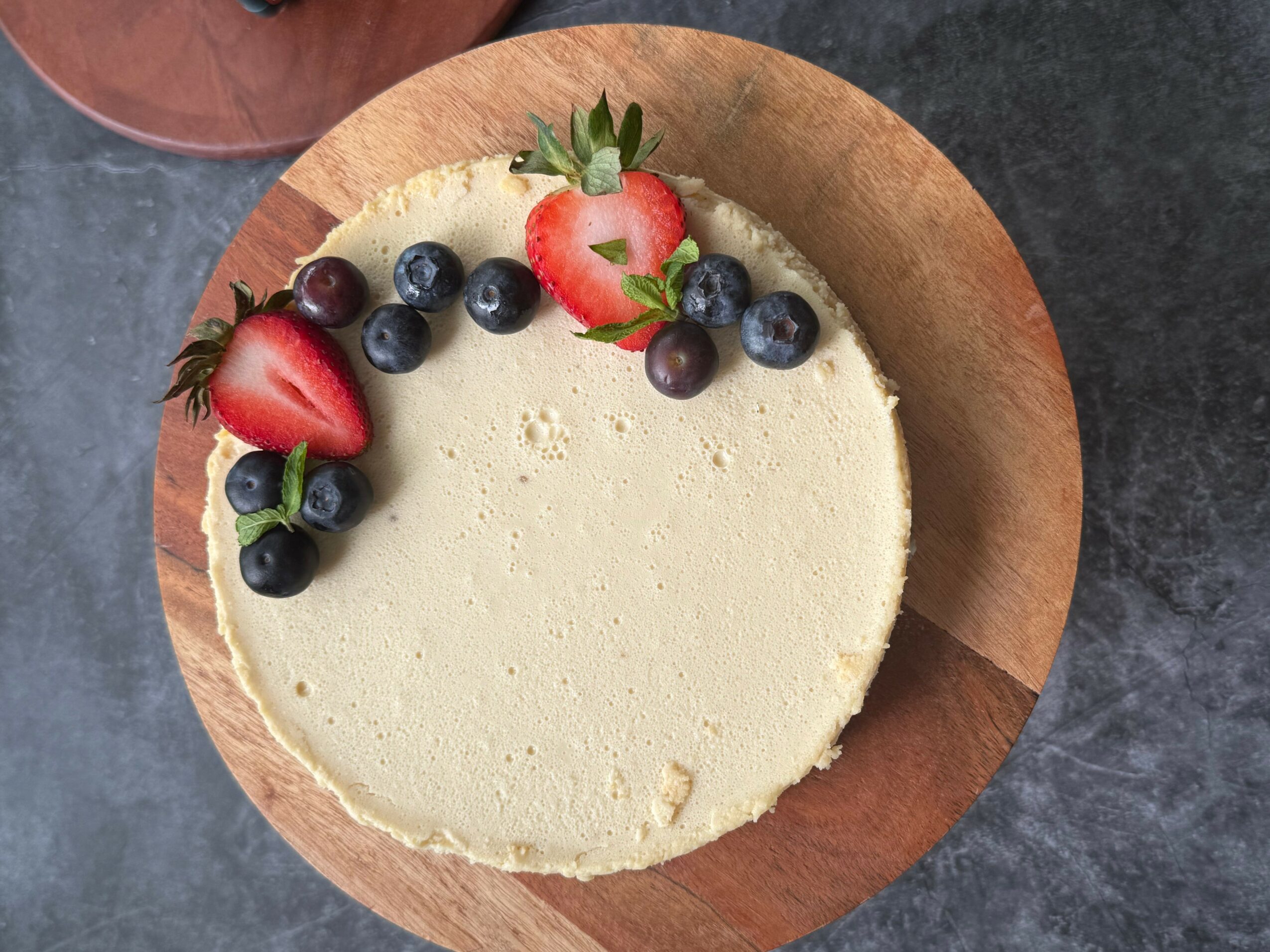 Round cheesecake topped with strawberries, blueberries, and grapes on a wooden plate, on a dark surface.