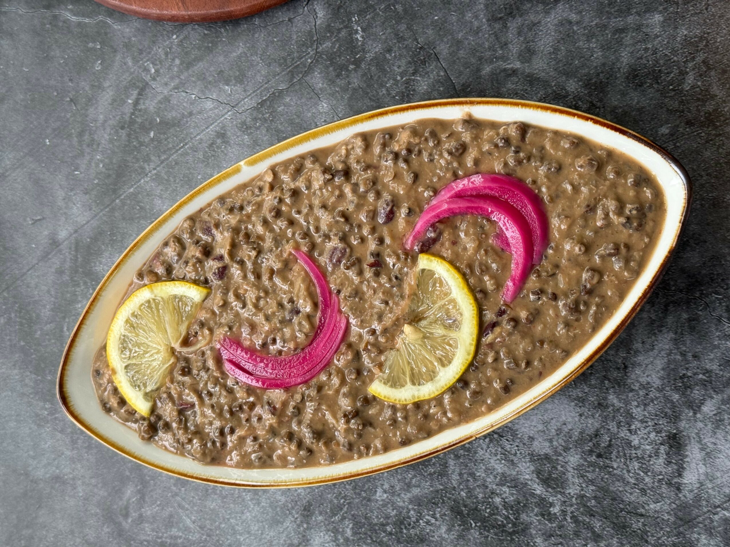 A dish of cooked lentils garnished with lemon wedges and pickled onions on a white oval plate, on a dark surface.