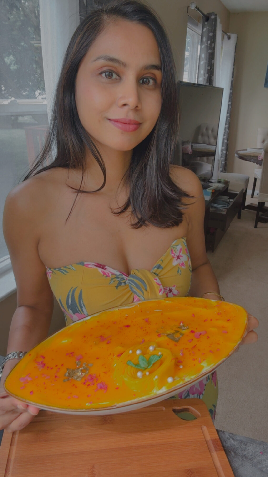 Woman holding a large colorful dish in a room with furniture and curtains.