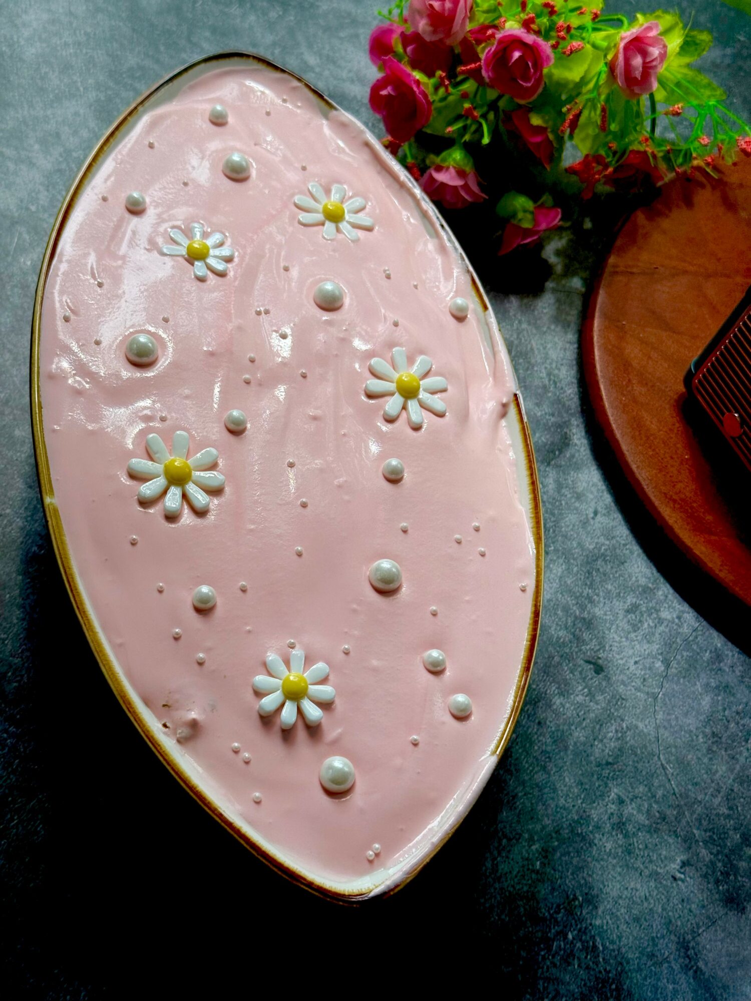 Oval cake with pink icing decorated with white daisies and small round decorations, placed on a dark surface.