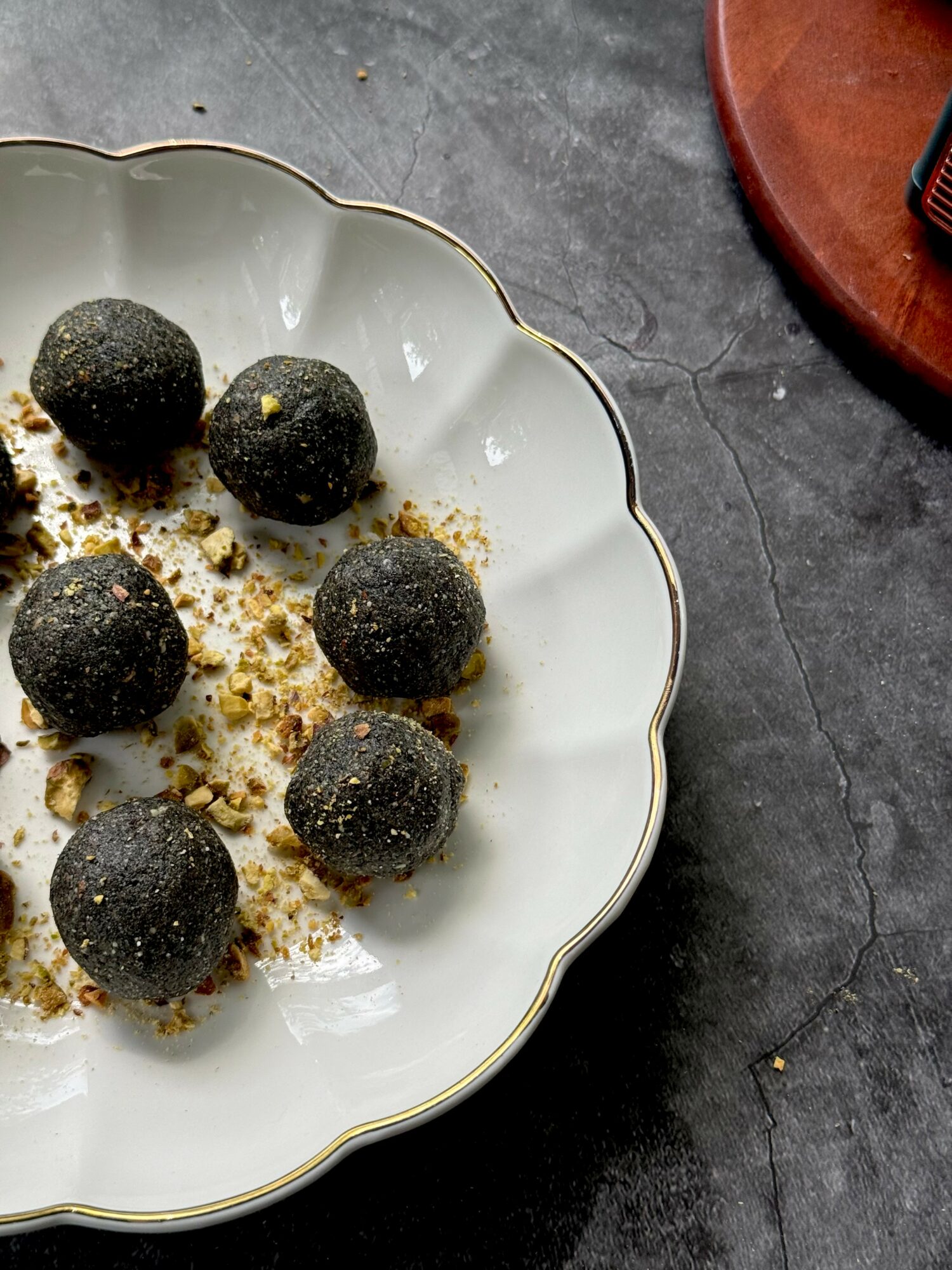 Six black spherical desserts on a white scalloped plate with crumbs, on a dark surface.