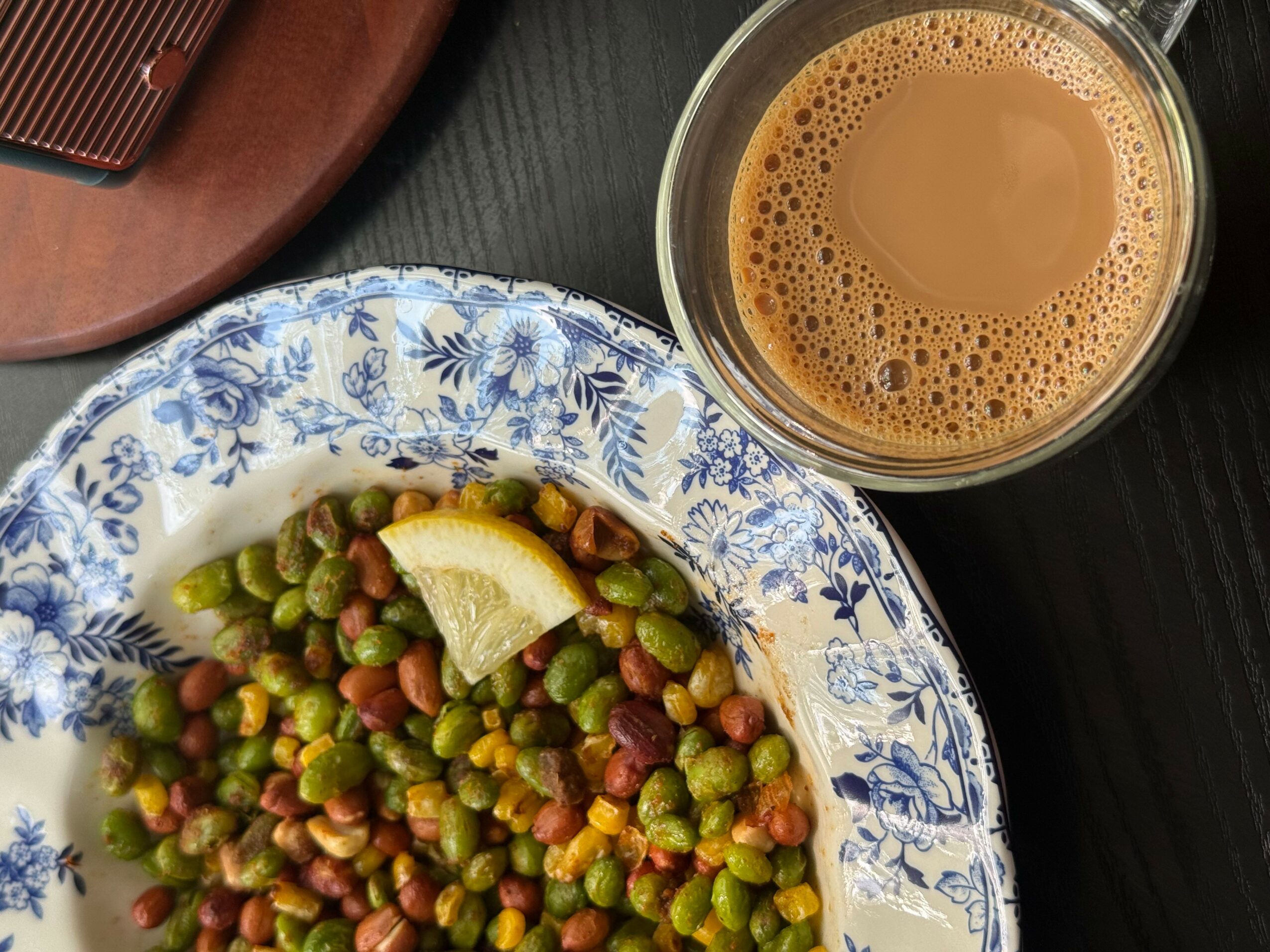 A bowl of mixed peas and beans with a lemon wedge, and a glass of coffee on a dark surface.