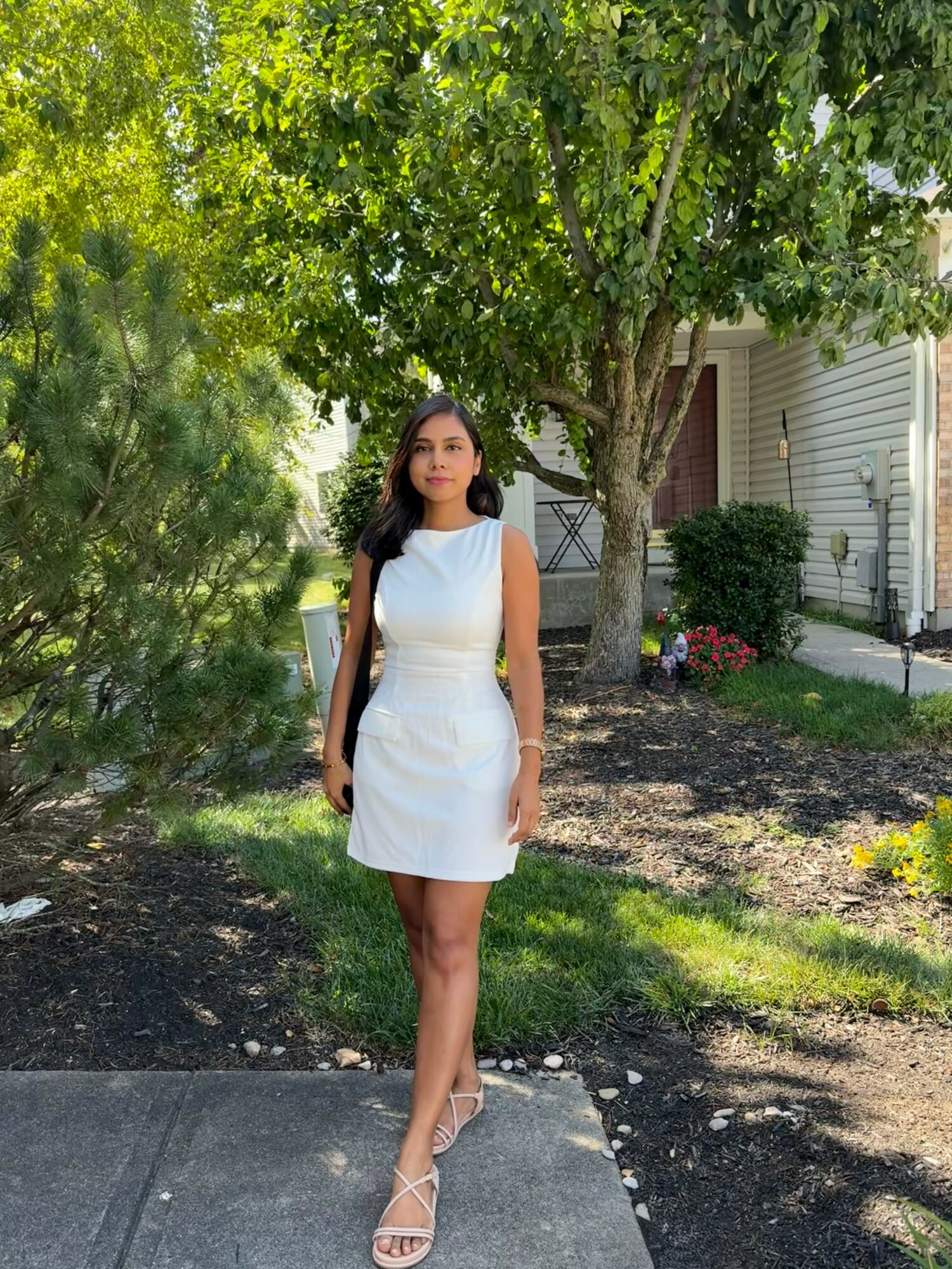 Young woman in white dress standing outdoors near trees and a house, sunlight filtering through leaves.