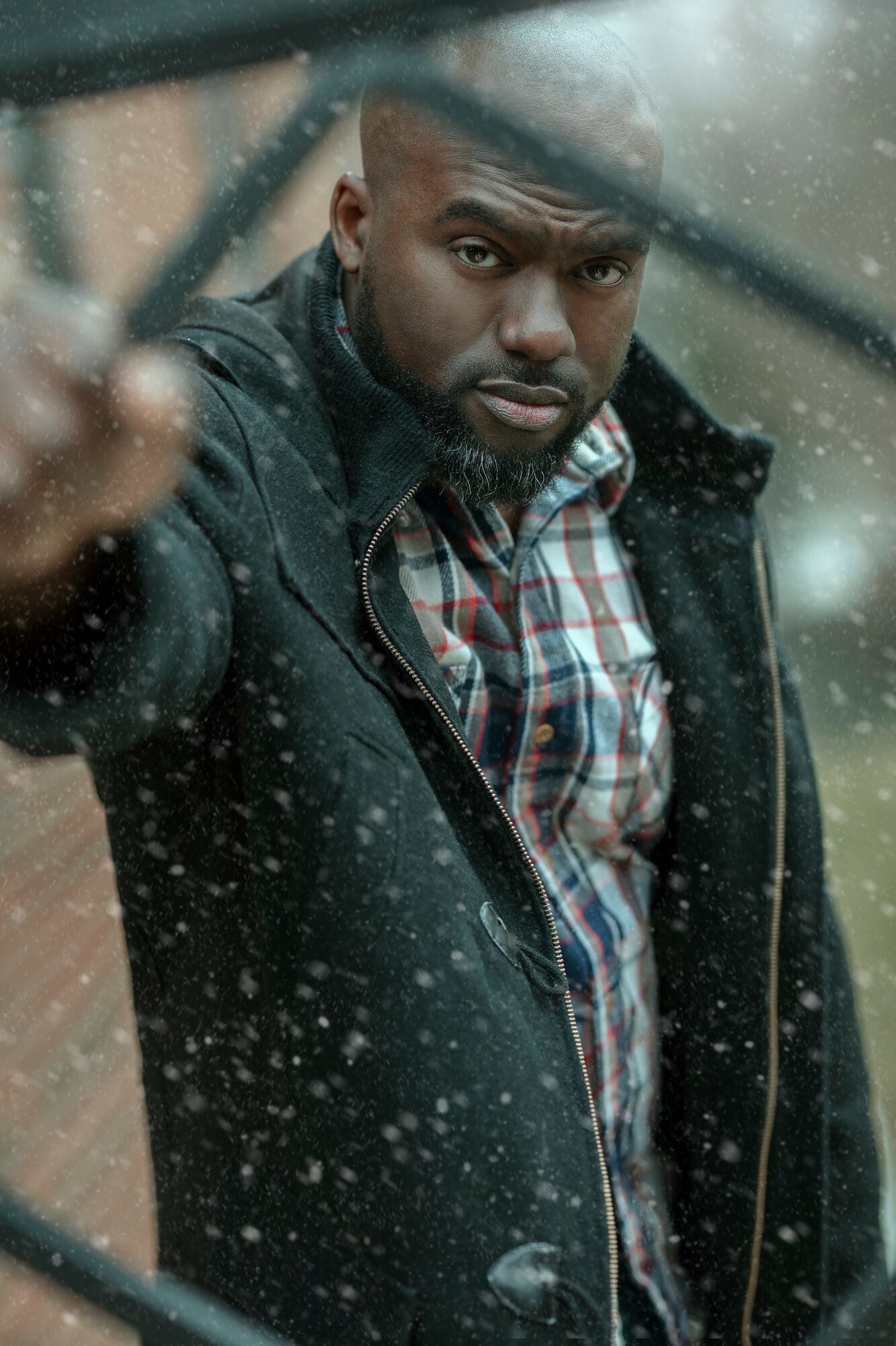 Man with beard and short hair looking through rain-covered window, wearing a plaid shirt and dark jacket.