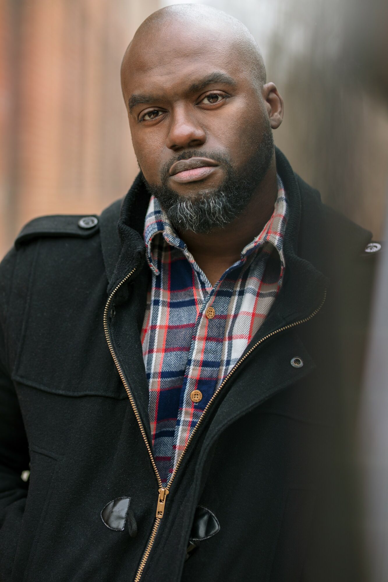 Man with a beard and serious expression wearing a plaid shirt and black jacket outdoors.