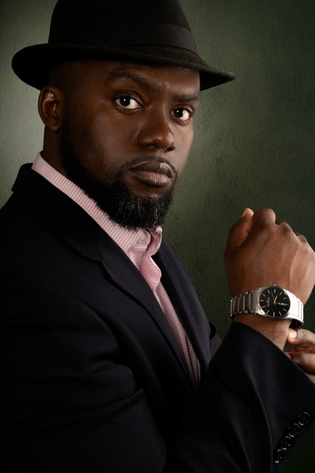 Man wearing a black hat and suit, with a pink shirt, looking serious, with a watch on his wrist, against a dark background.