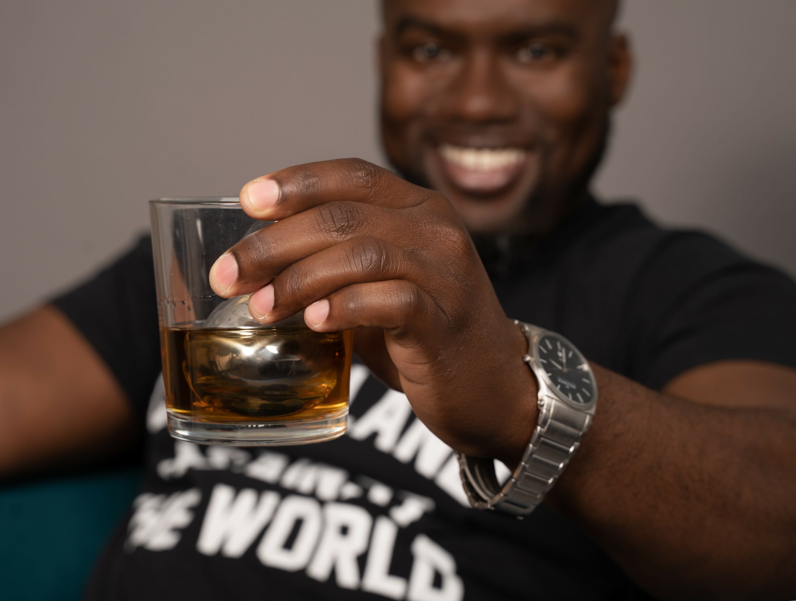 Person holding a glass of dark beverage, smiling, wearing a watch and a black shirt, against a plain background.