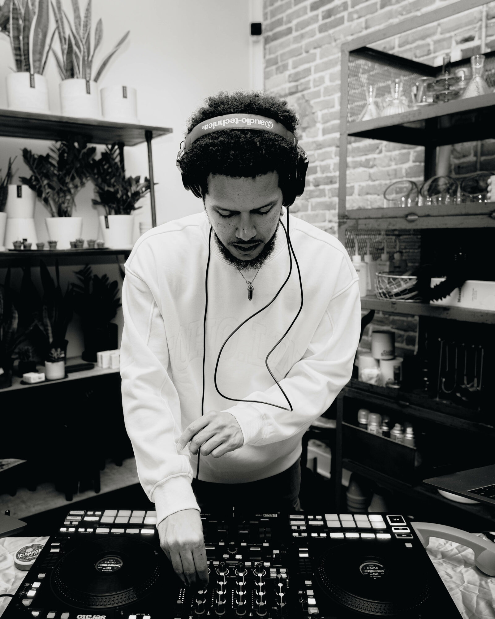 Person with headphones operating a DJ controller in a room with shelves and plants.