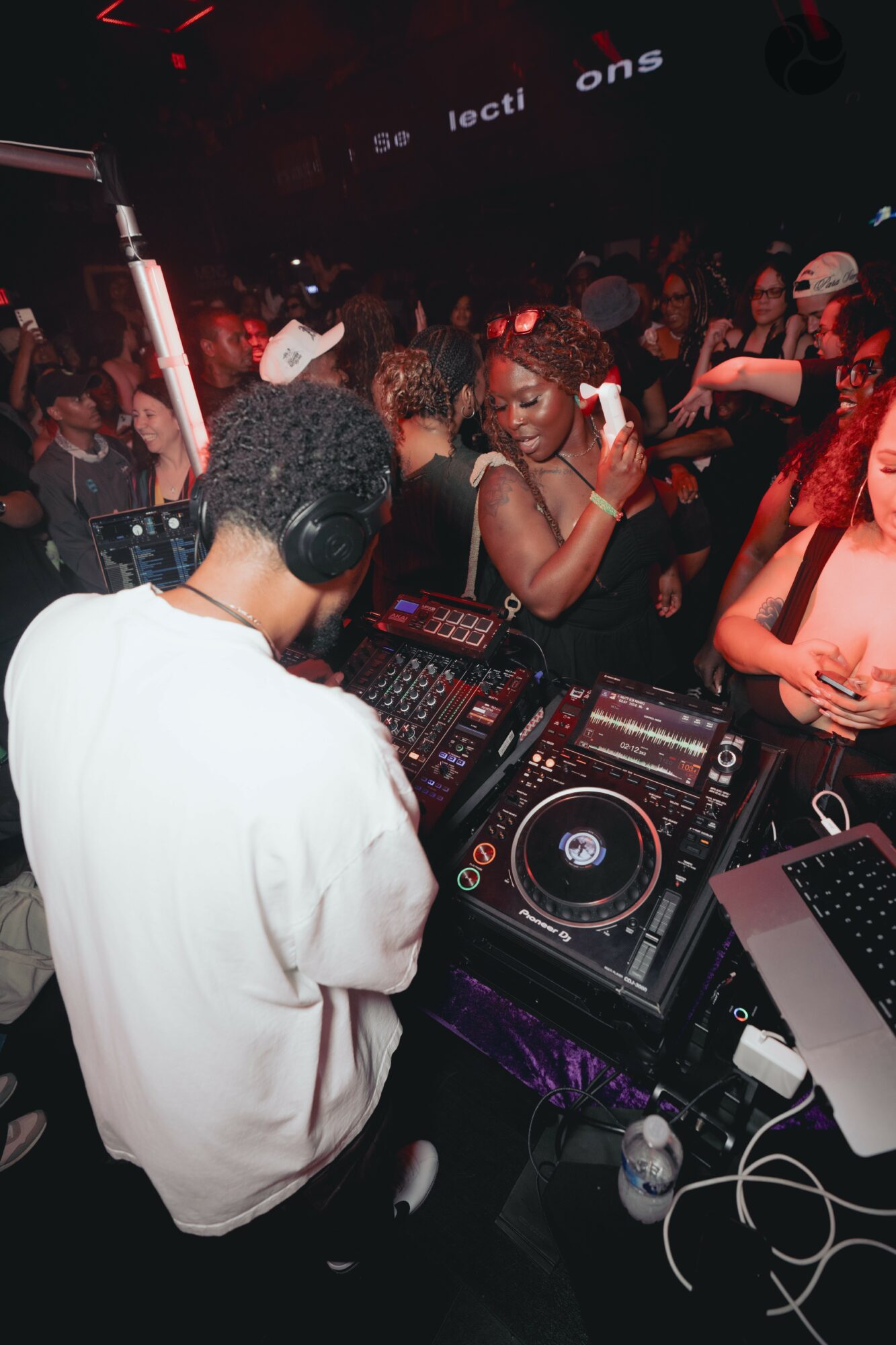 DJ with headphones at turntables in a crowded nightclub with dancing people, dark lighting, and red accents.