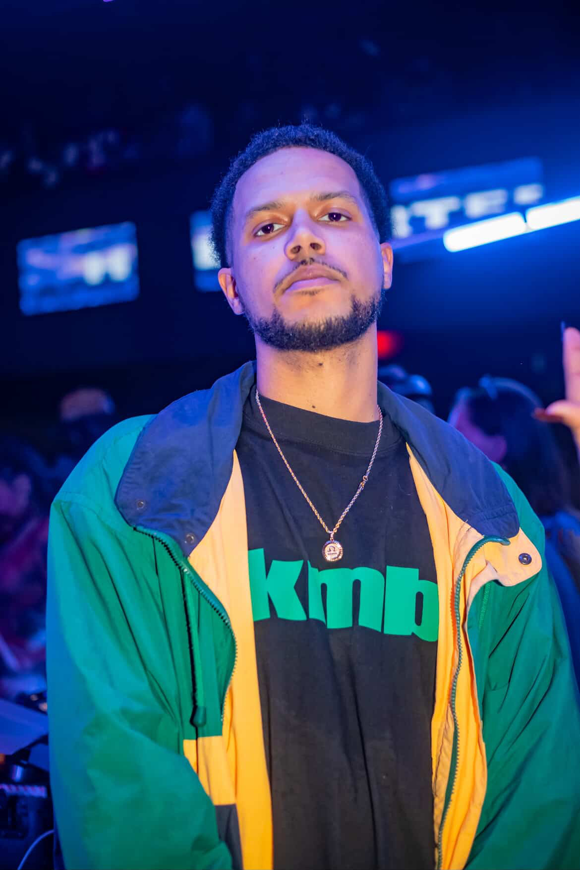 Young man with short hair and beard in a colorful jacket and black shirt, standing in a dimly lit environment with blue lighting.