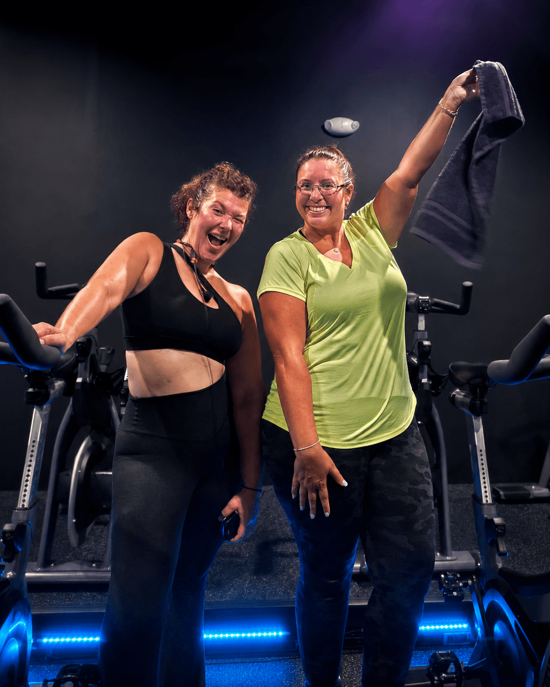 Two women smiling and celebrating on exercise bikes in a gym, one holding a towel in the air.