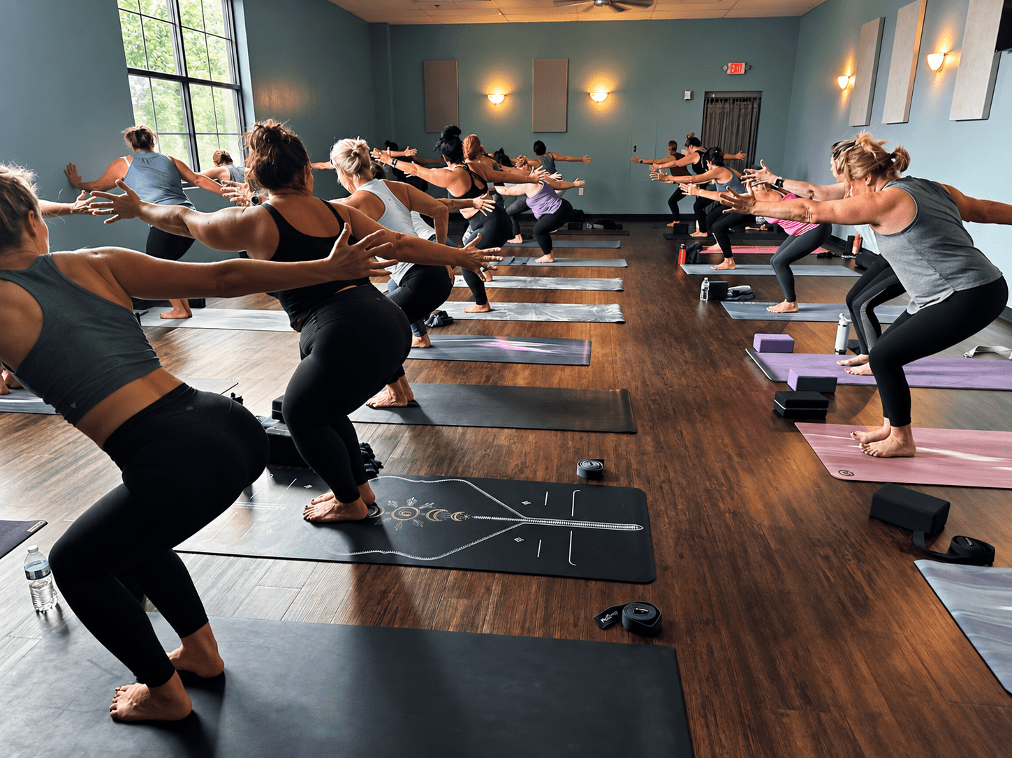 Group of people practicing yoga in a spacious room with large windows, wooden floor, and blue walls, all in a squatting pose.