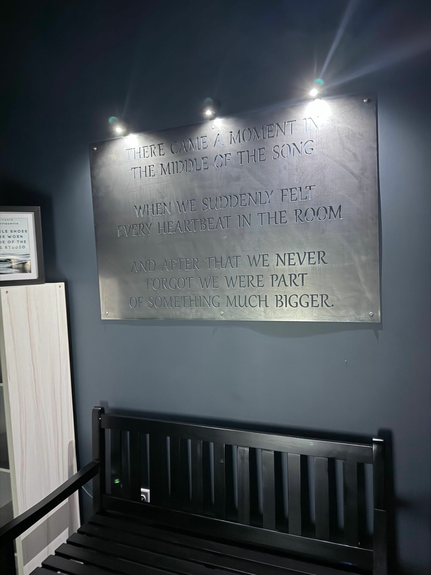 Metal plaque with engraved text mounted on a wall, illuminated by bright lights, with a black slatted object nearby.