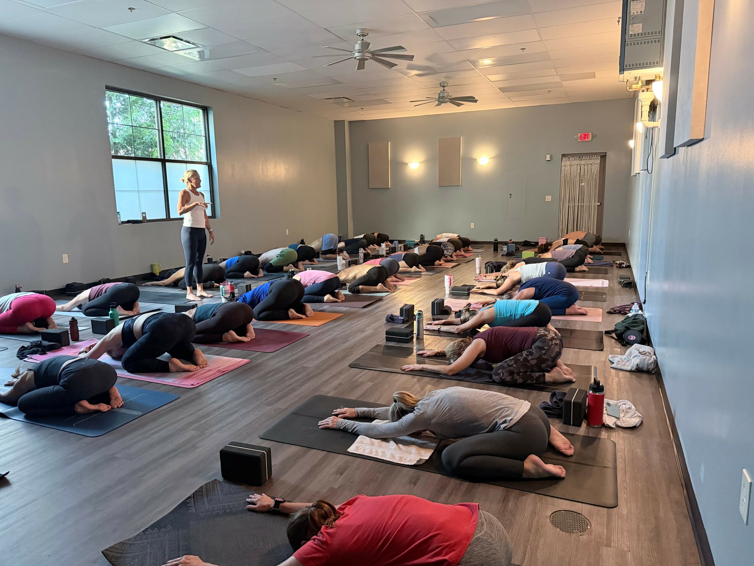 People practicing yoga in a spacious room with large windows, lying on mats in a forward bend pose, instructor at front.