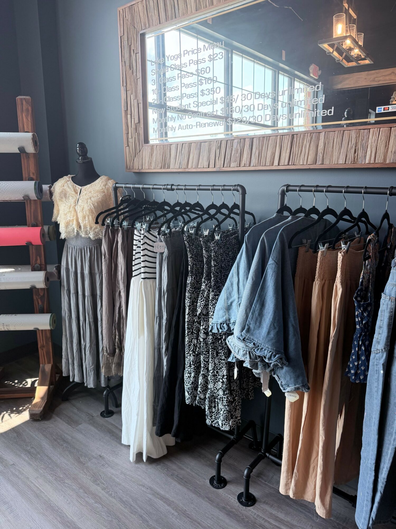 Clothing racks with various garments in a store, window with a view outside, wooden wall, and gray flooring.