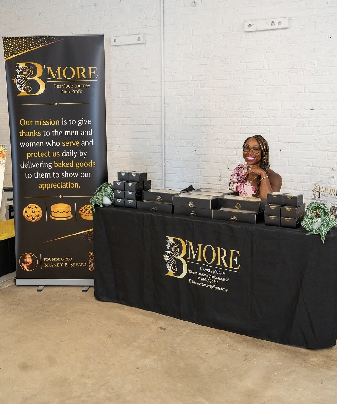A smiling woman sits behind a table with baked goods at a promotional event, with a banner displaying '3 MORE' and a mission statement.