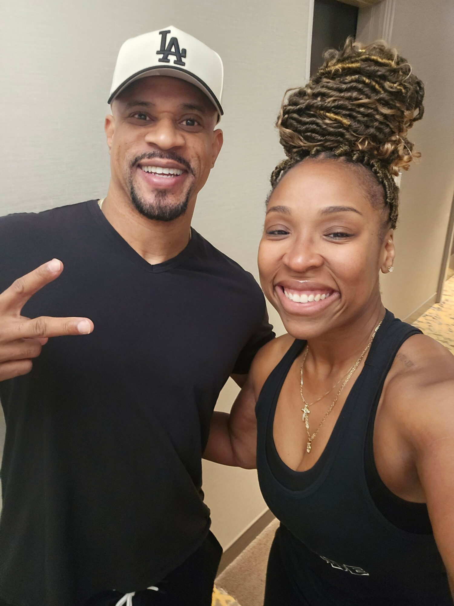 Two people smiling, one wearing a black tank top and the other wearing a black shirt and a white cap, indoors.