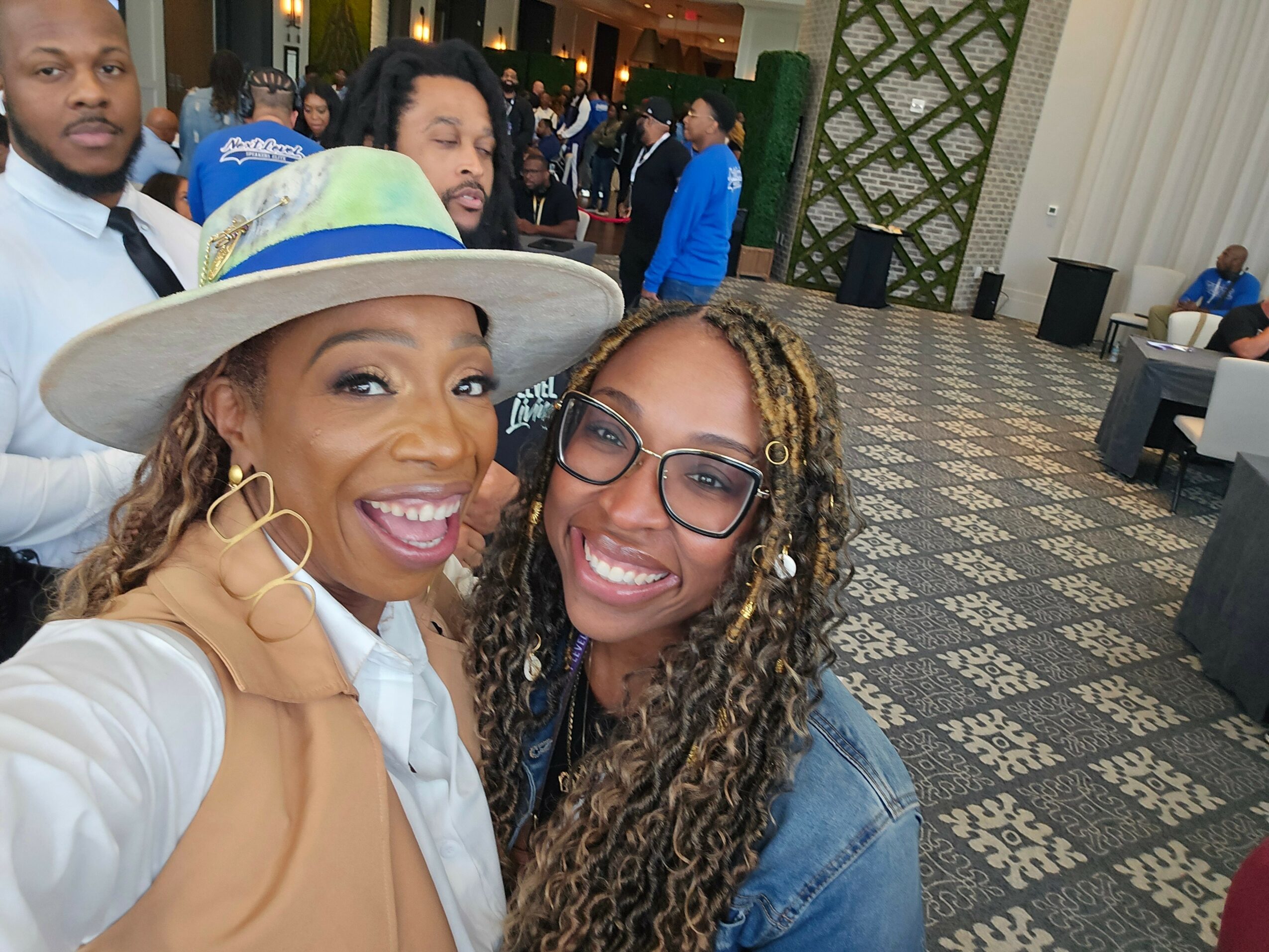 Two women smiling, one wearing a large hat and white blazer, the other with glasses and curly hair, in a decorated indoor setting.