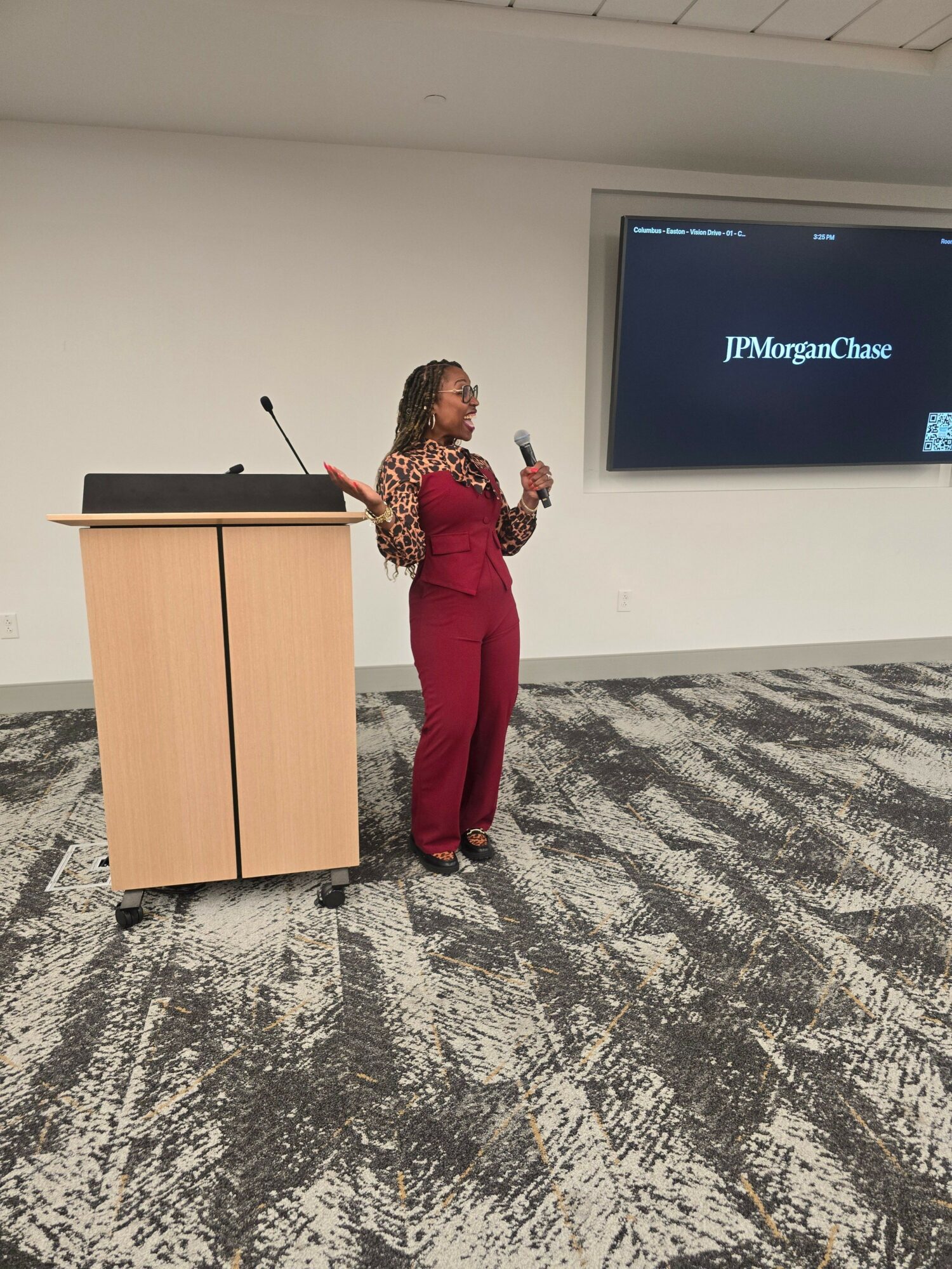 Woman standing next to a wooden podium in a conference room, with a screen displaying a logo behind her.